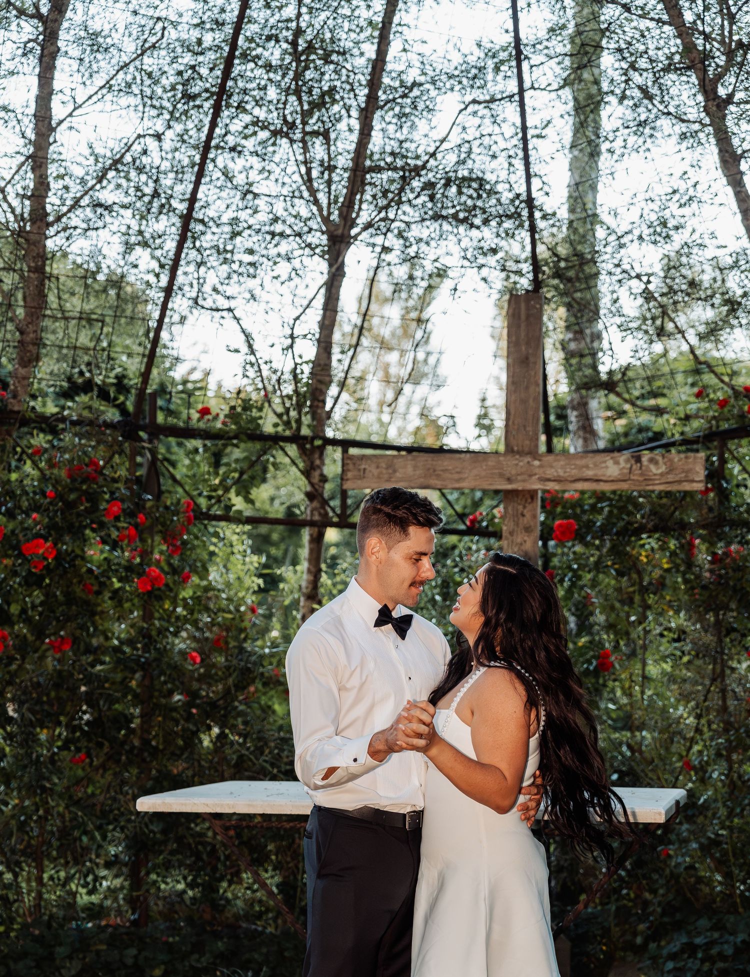 Bride and groom share a tender moment beneath a rustic wooden arch adorned with red roses outdoors.