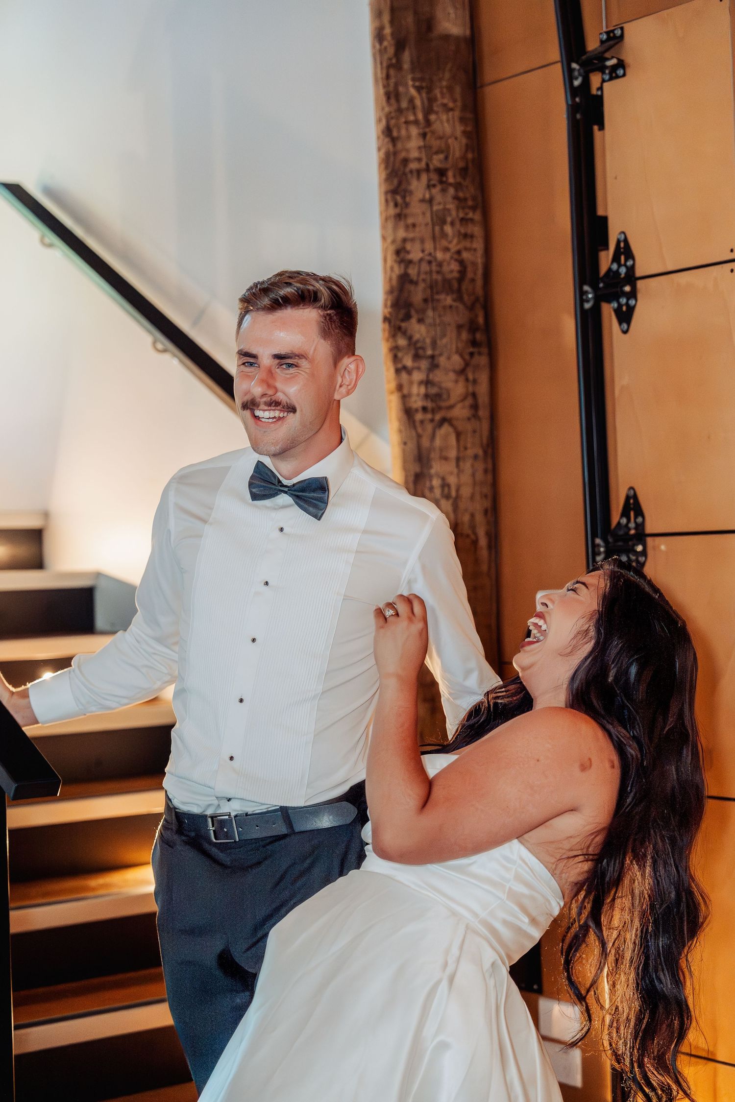 Laughing bride in strapless gown playfully grabs groom in bow tie near rustic wooden staircase indoors.