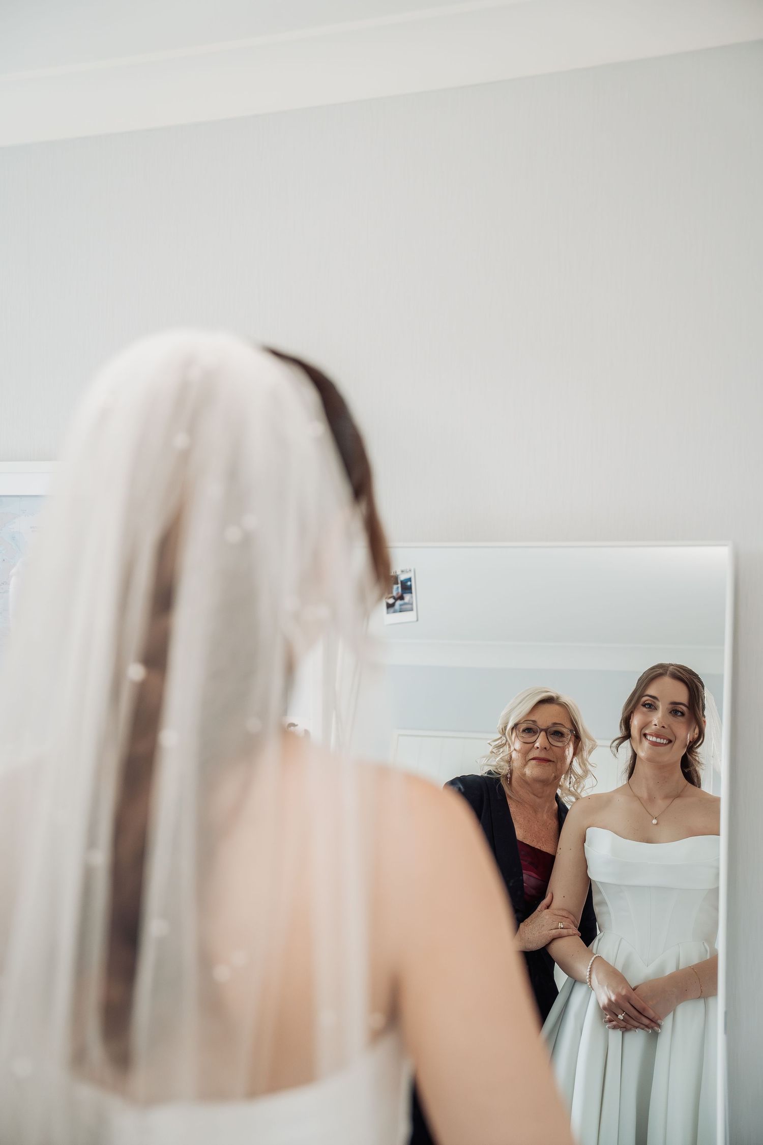 Bride in white strapless gown and veil smiles at her reflection as an older woman helps her prepare.