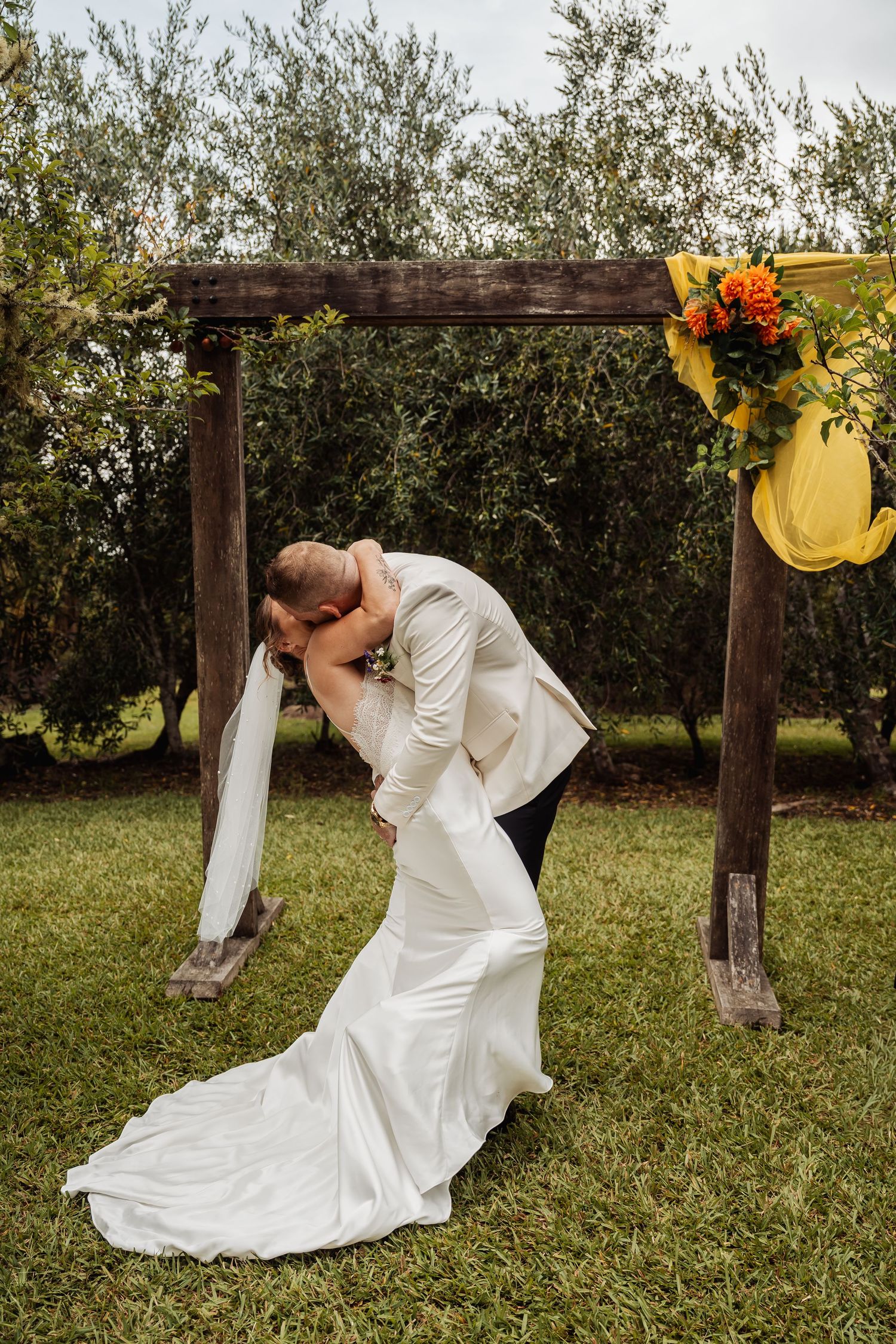 Bride and groom share a romantic dip kiss under a rustic wooden arch adorned with colorful flowers at an outdoor wedding.