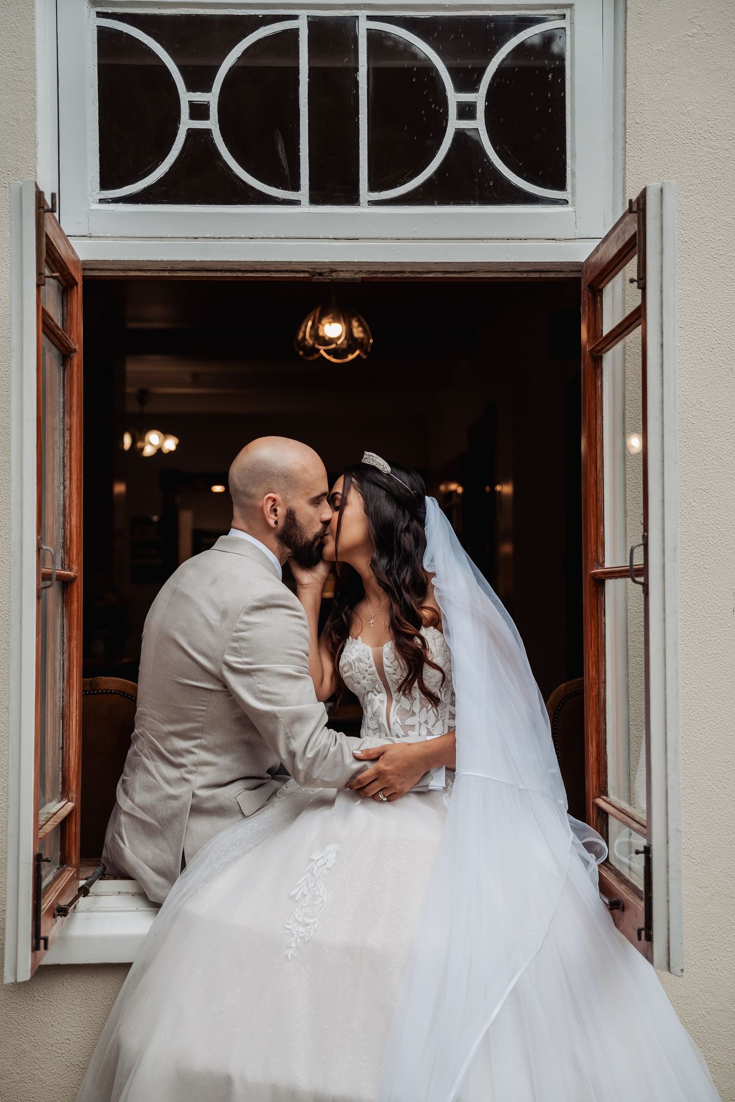 Bride and groom sharing a kiss in an open window, bride in white gown with veil.