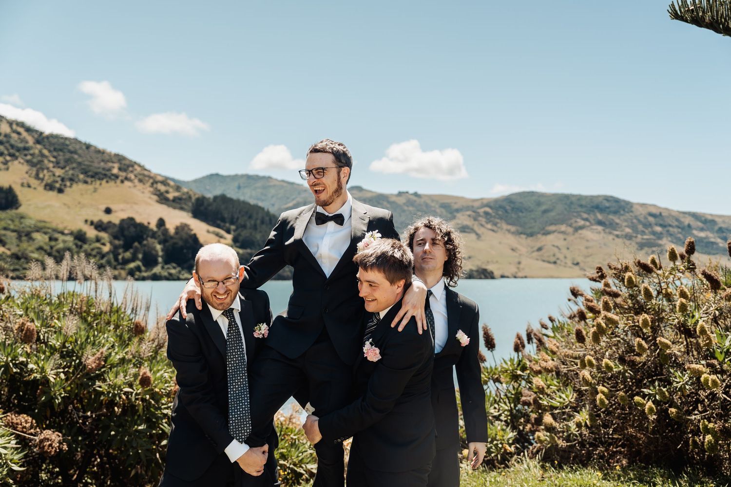 Groom and groomsmen posing playfully outdoors with scenic lake and mountains in background on wedding day.