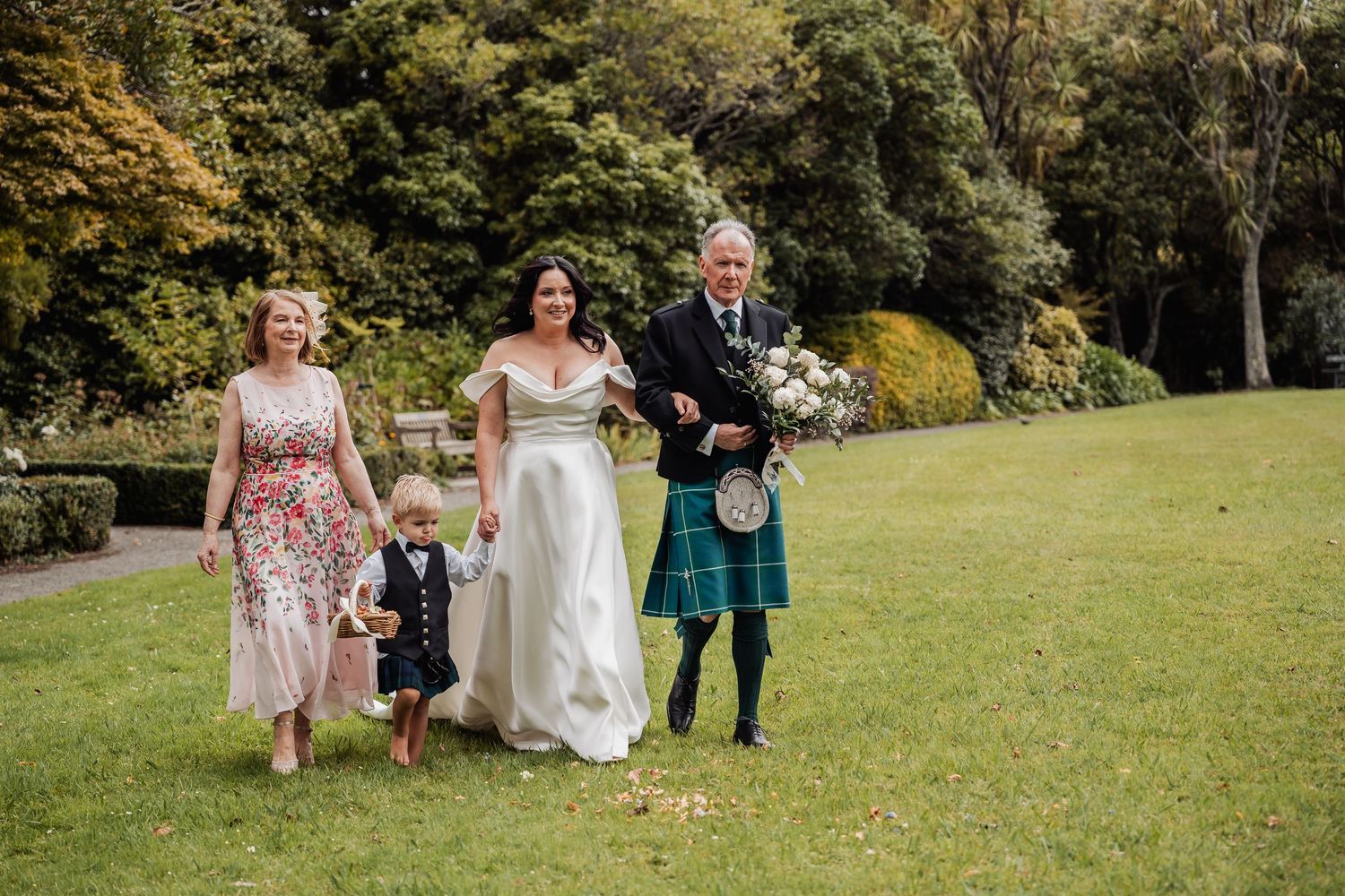 Bride in white gown walks with family on green lawn, man in blue tartan kilt holding flowers at Scottish wedding.