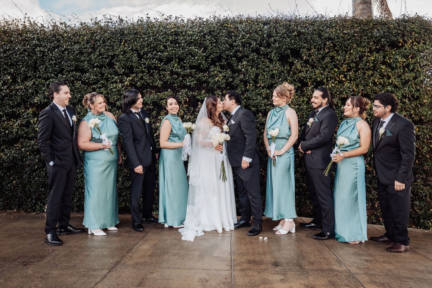 Wedding party in turquoise dresses and black suits lined up outdoors against greenery backdrop.