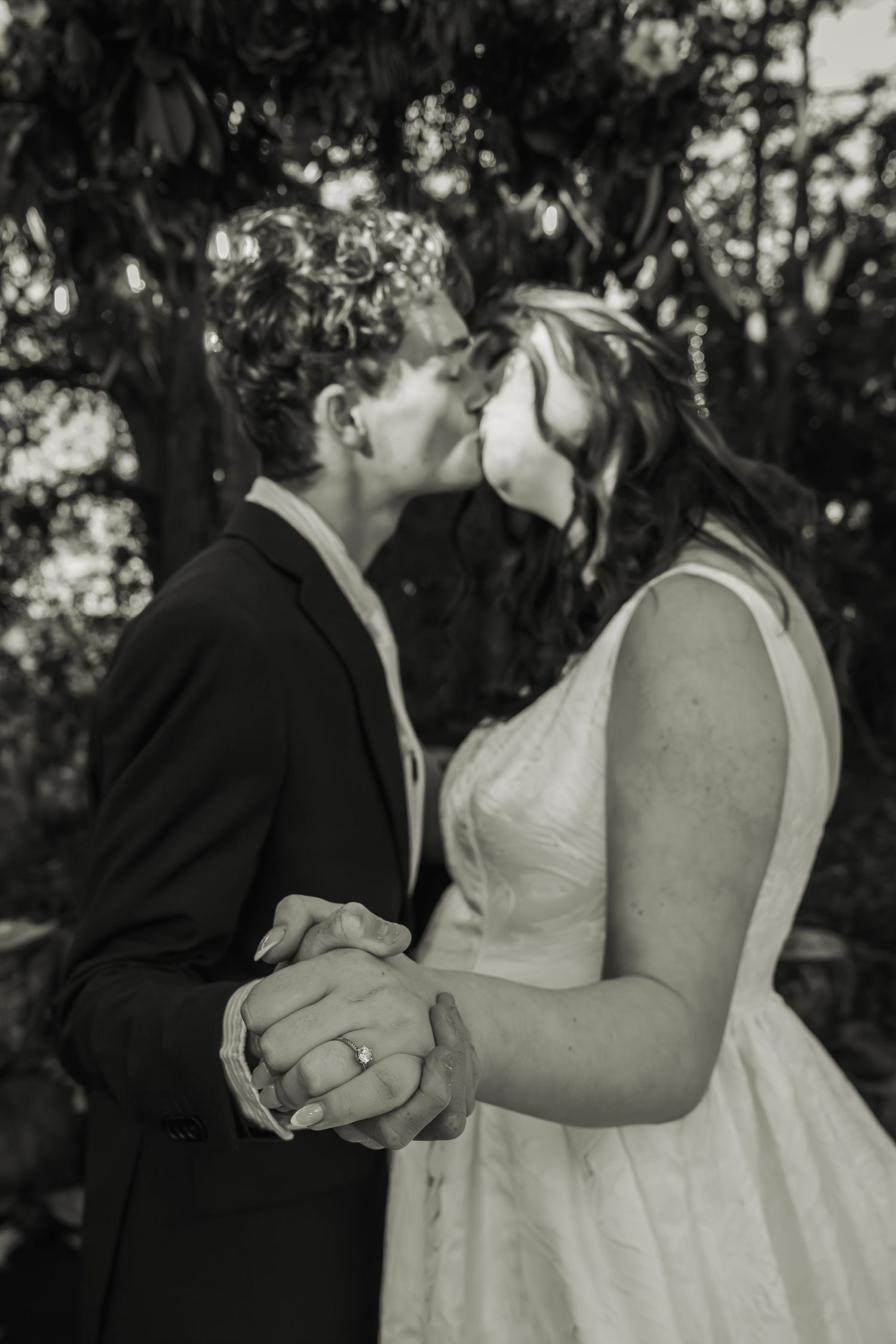 Black and white romantic photo of newlyweds kissing while holding hands in garden setting.