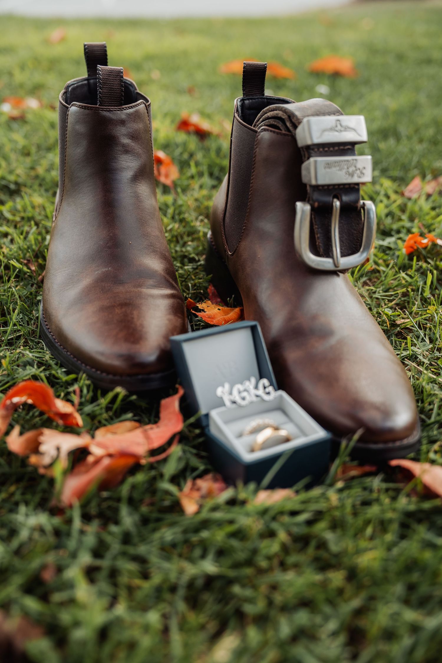 Brown leather Chelsea boots with a belt and wedding ring box on autumn grass.