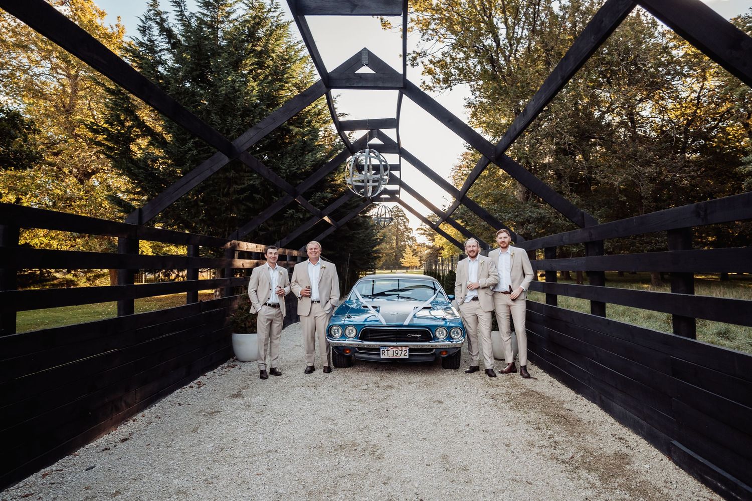 Groomsmen in tan suits posing with blue muscle car on bridge.