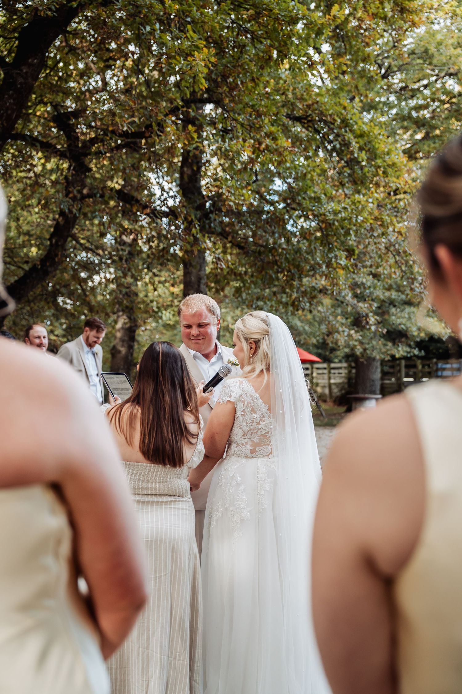 Bride in lace wedding gown exchanges vows with groom outdoors under large trees.