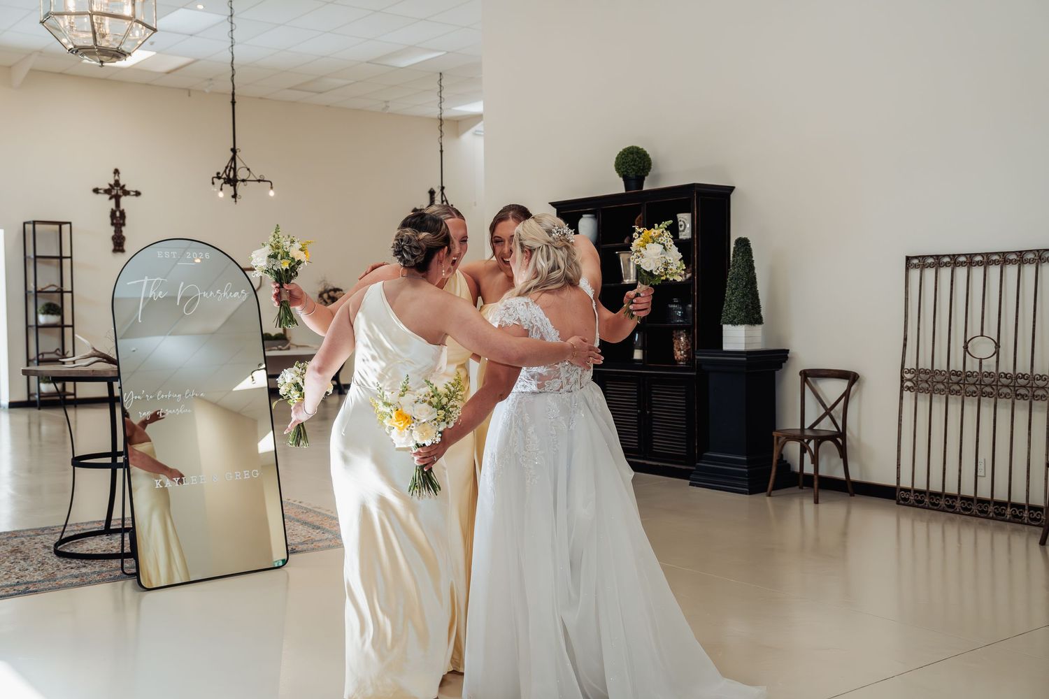 Bride in lace gown and bridesmaids with bouquets sharing a joyful moment near a mirror in a bright bridal suite.