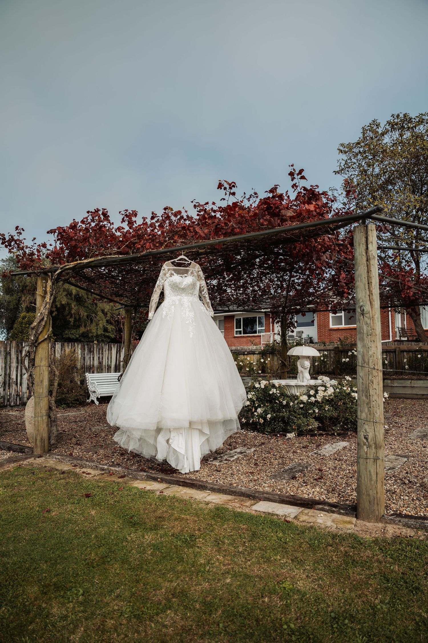 Elegant white ball gown wedding dress hanging on wooden pergola in garden with autumn red trees.