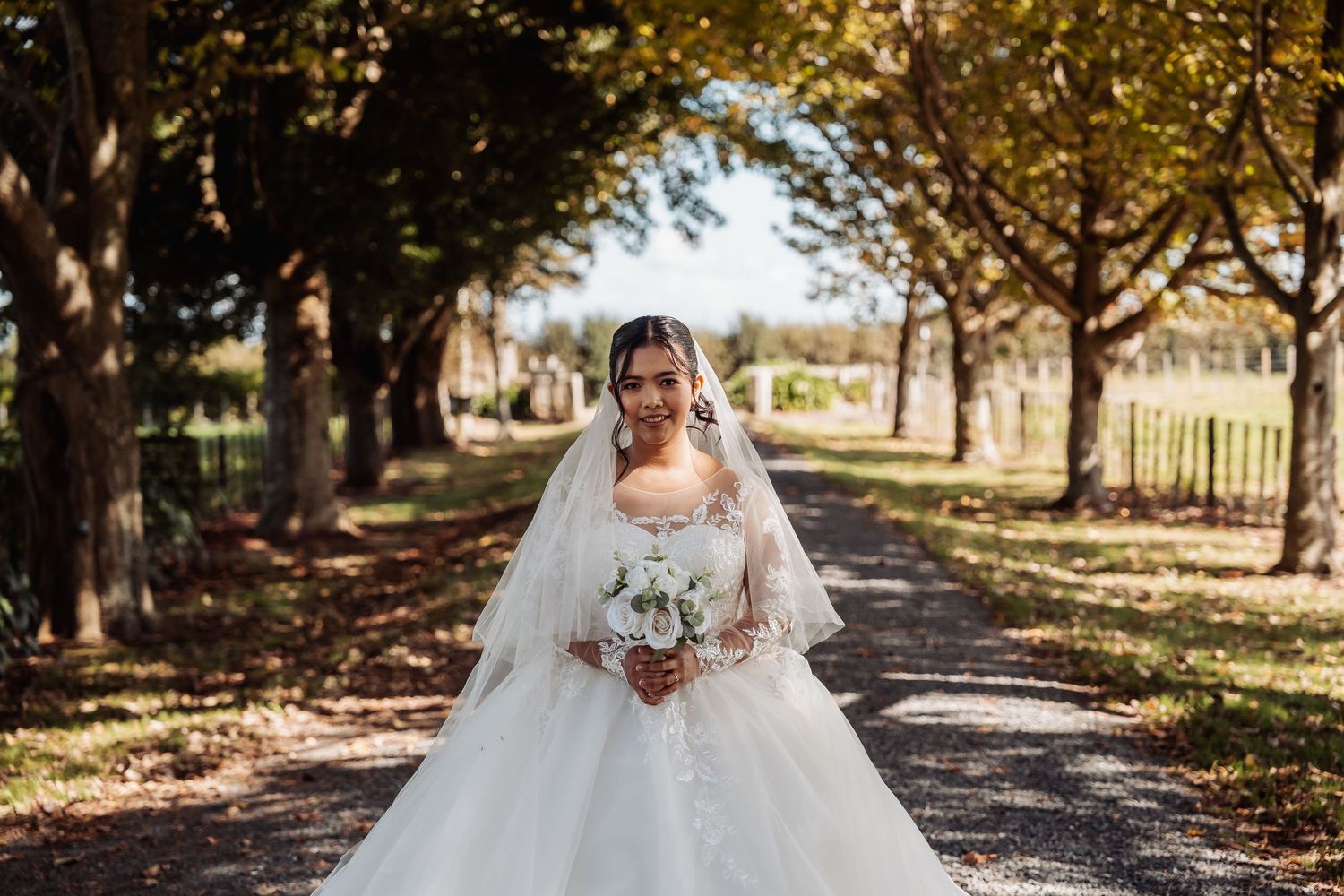 Bride in a white ball gown holding a bouquet walking down a tree-lined path on her wedding day.