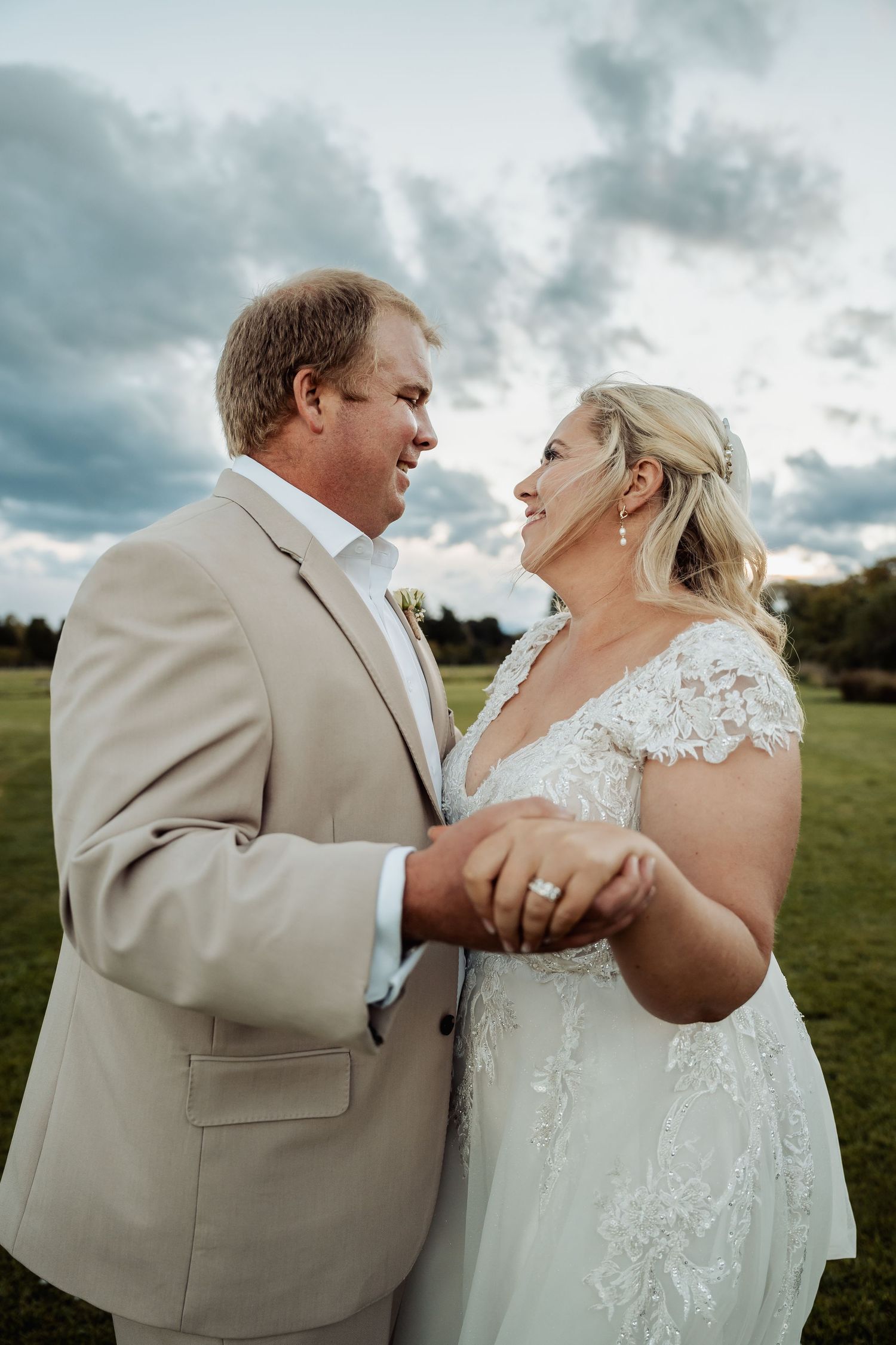 Bride and groom hold hands and gaze at each other lovingly under a dramatic cloudy sky.