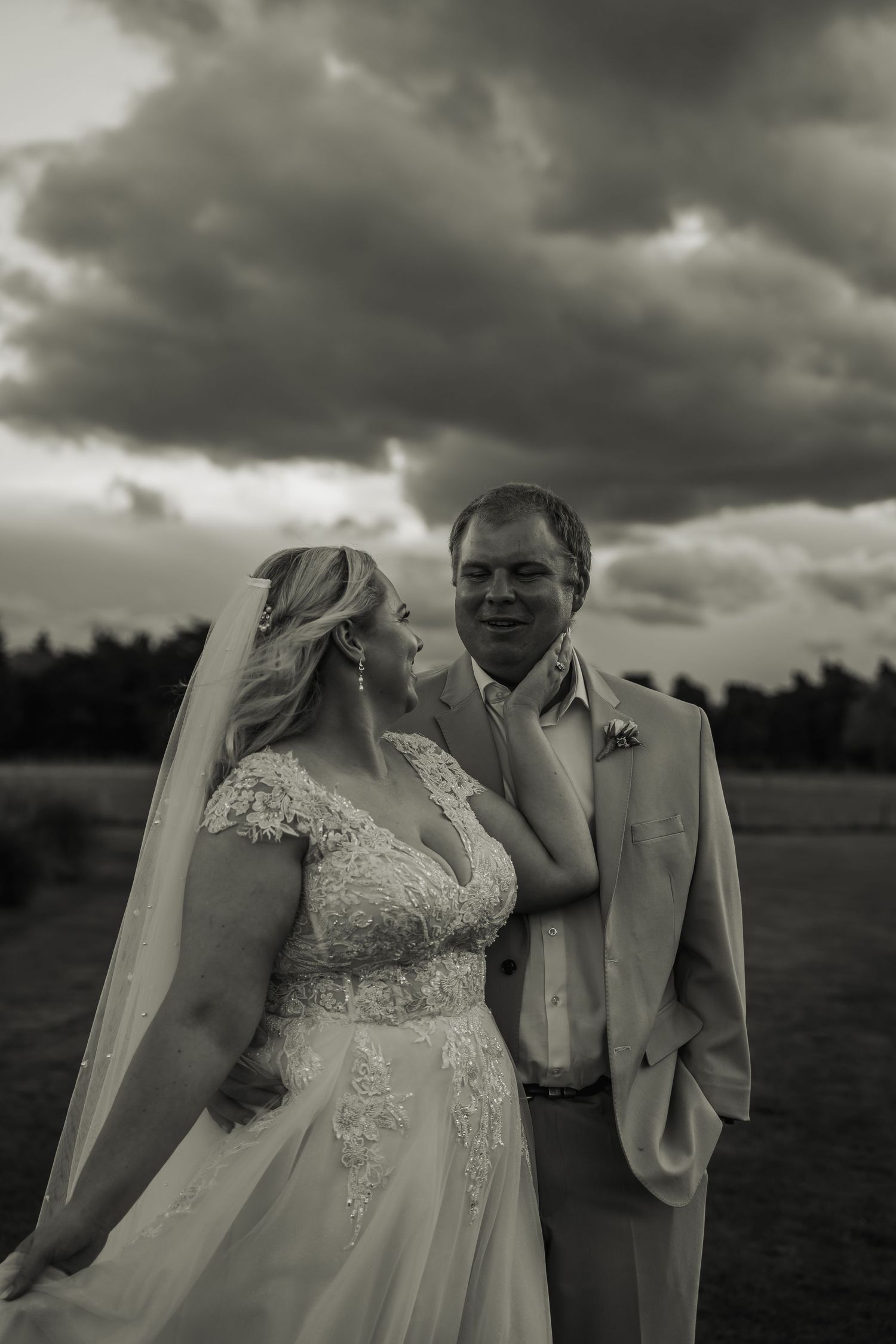Black and white portrait of bride in lace veil gown and groom in tan suit under dramatic clouds.