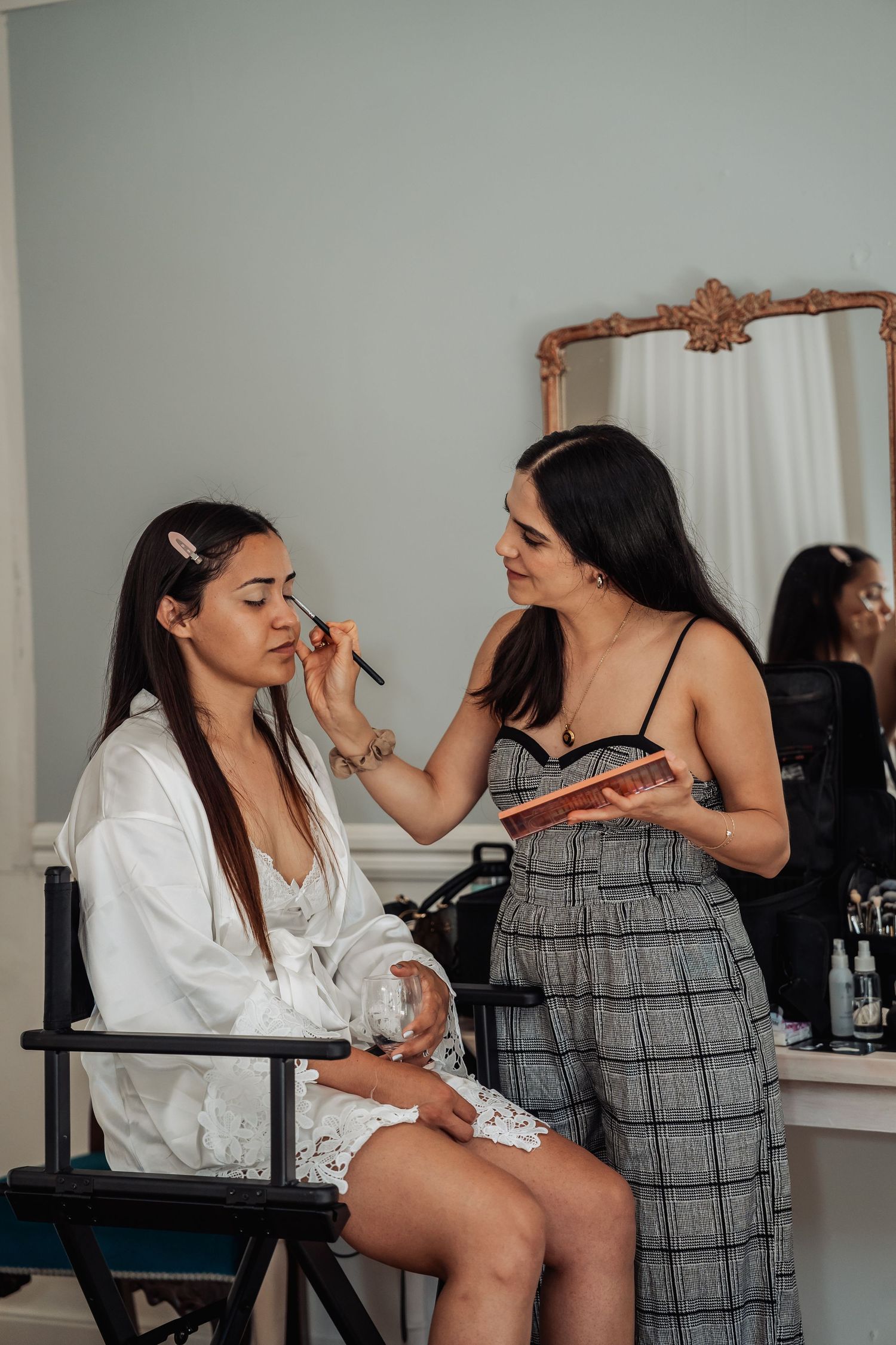 Makeup artist applying eyeshadow to bride in white robe seated in director chair before wedding.