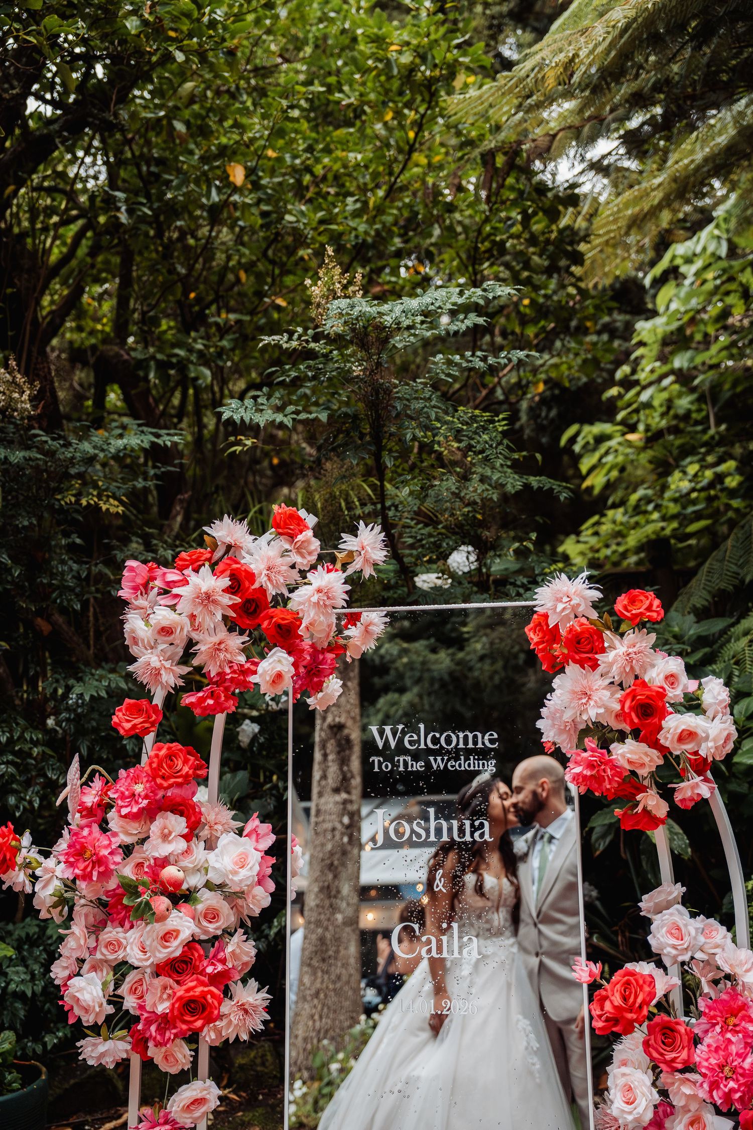 Couple kissing under a floral arch with pink and red flowers at Joshua and Caila wedding ceremony outdoors.