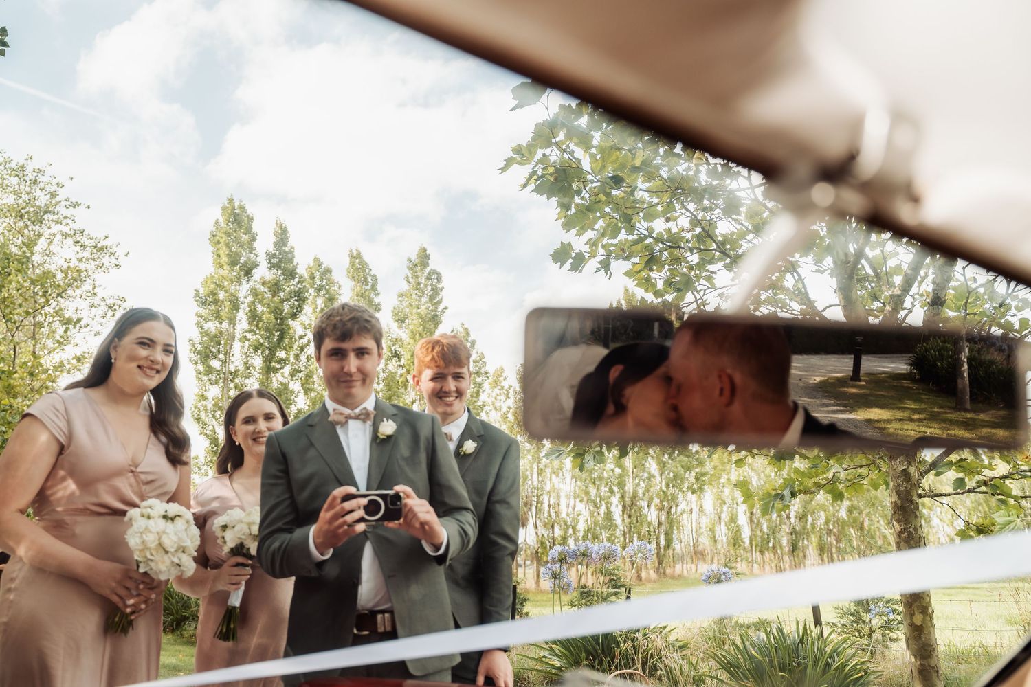 Groom holds camera while bridal party stands outdoors, with kissing couple reflected in vintage car mirror.