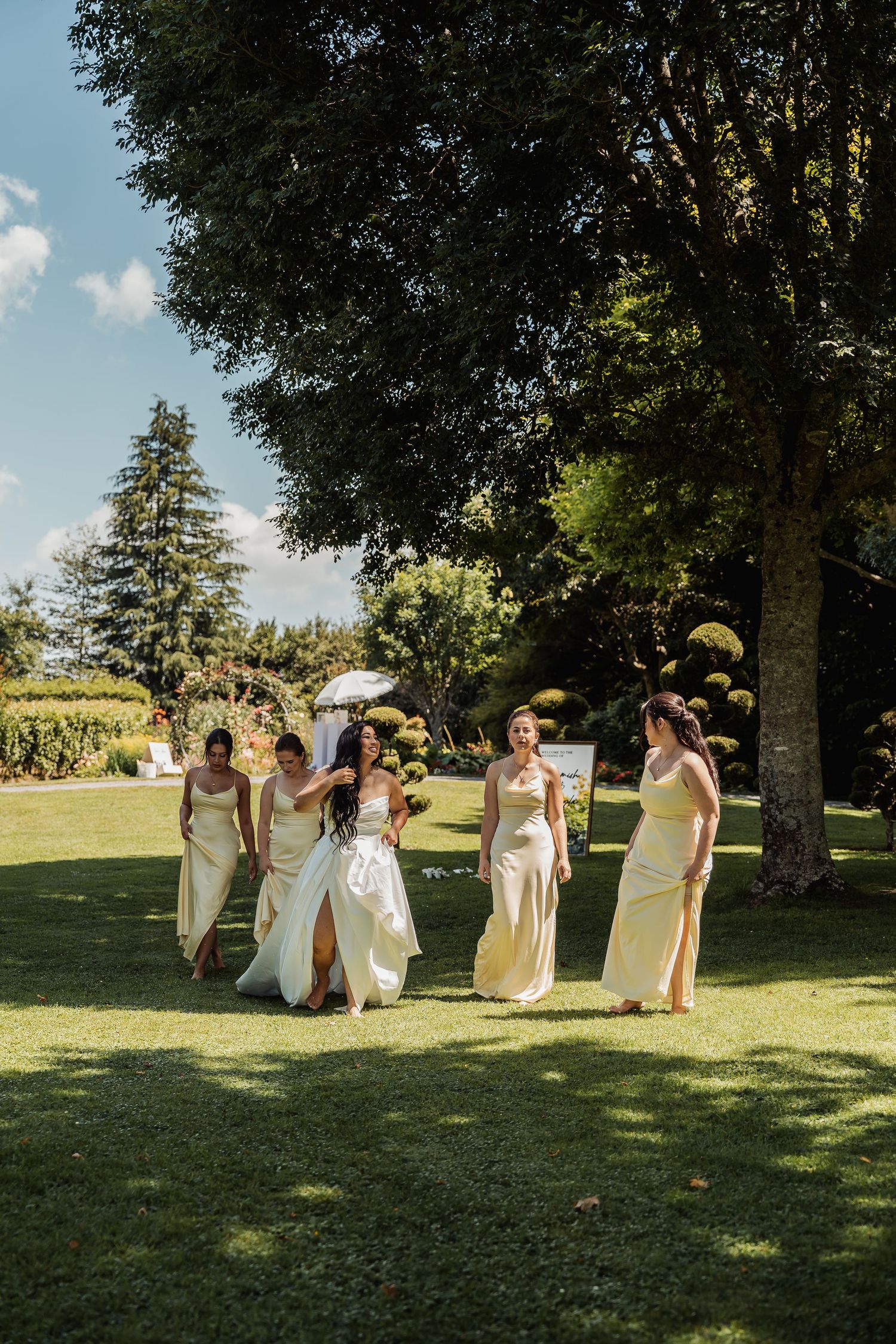 Bride in white gown walks with four bridesmaids in yellow dresses across lush green garden on sunny wedding day.