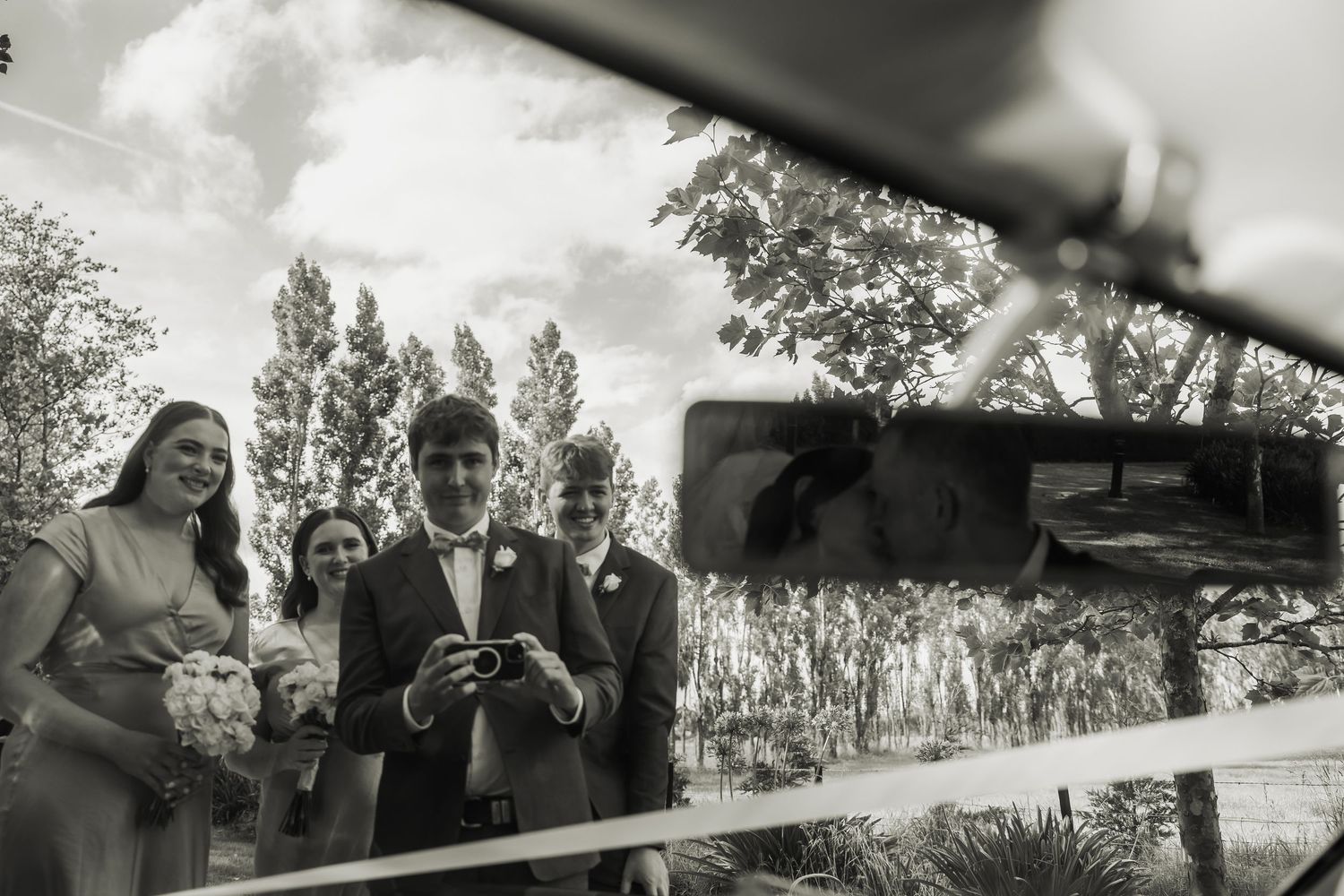 Black and white reflection of groom and wedding guests captured in car mirror outdoors.