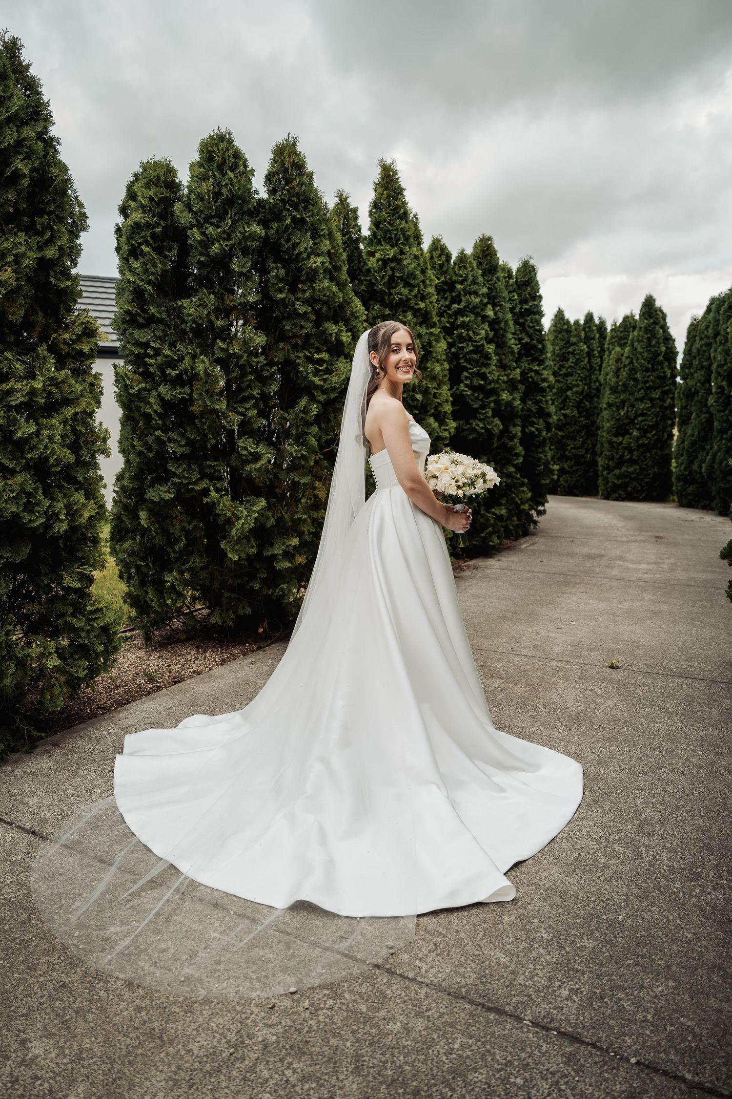 Bride in elegant white gown with long train and veil holds bouquet beside tall evergreen trees.