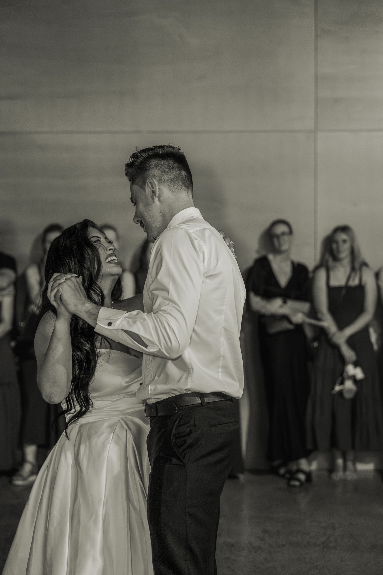 Black and white photo of bride and groom sharing an emotional first dance with wedding guests watching in background.