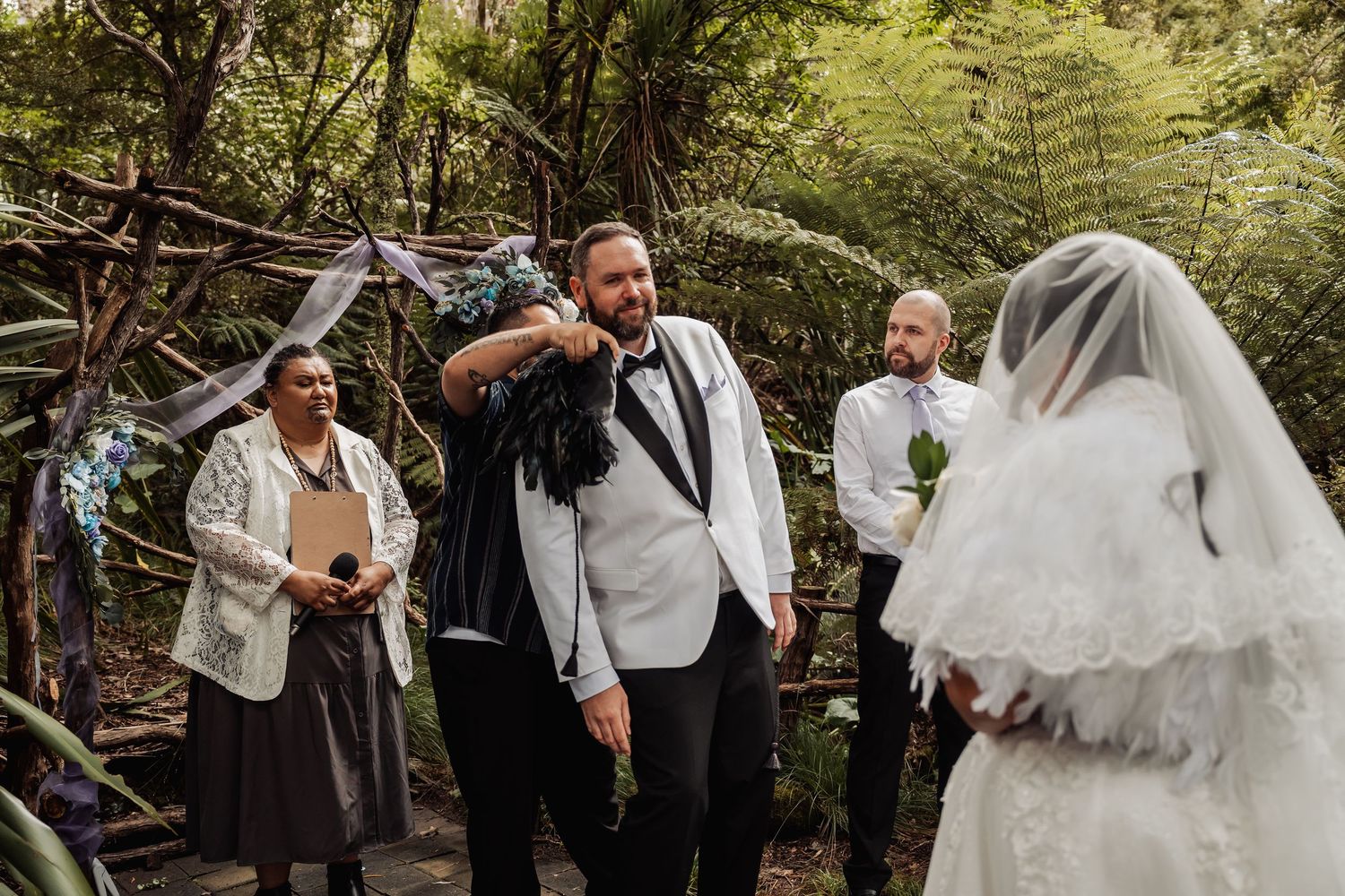 Groom in white tuxedo with Maori feather cloak reacts emotionally as bride approaches forest ceremony altar.