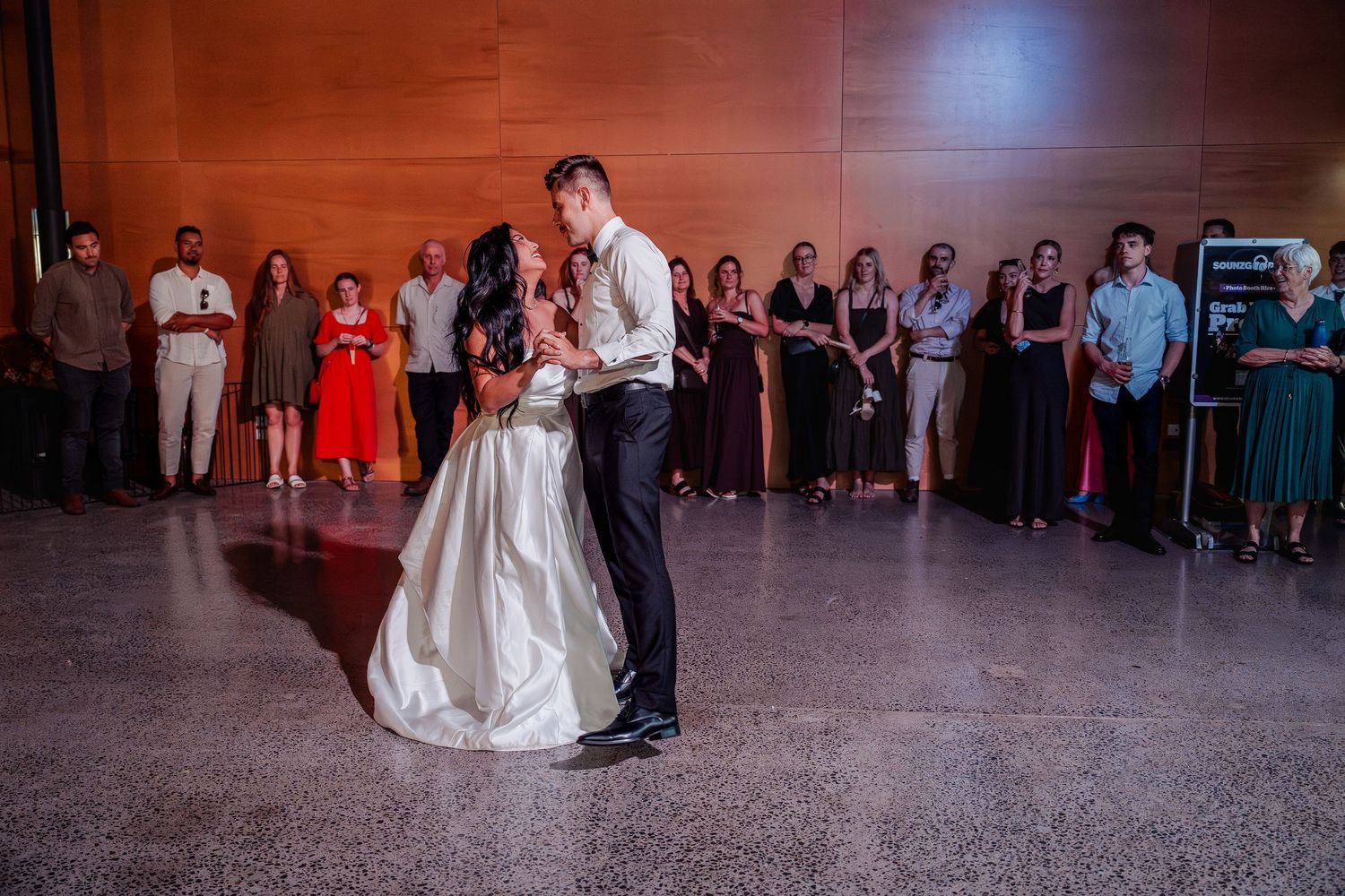 Bride and groom share romantic first dance while wedding guests watch in elegant indoor venue.
