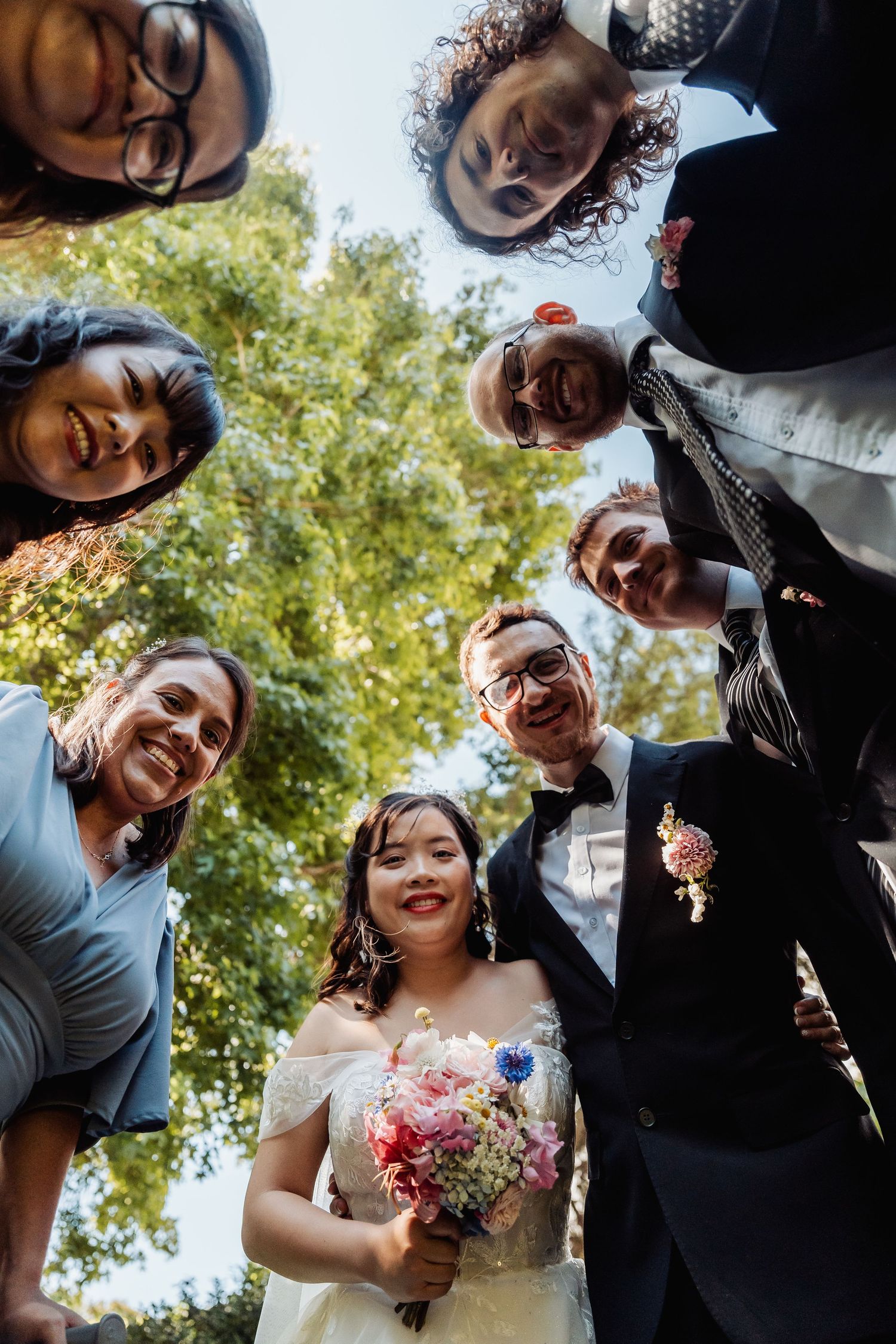 Wedding party posing for fun low-angle group photo with bride and groom in center outdoors.