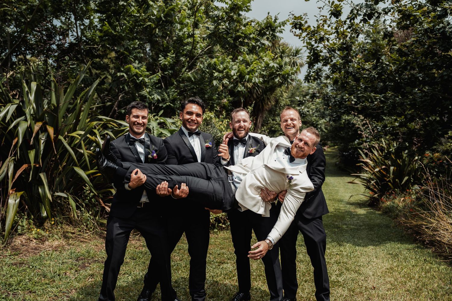 Four groomsmen in black tuxedos lifting groom in white jacket during fun outdoor wedding photo.