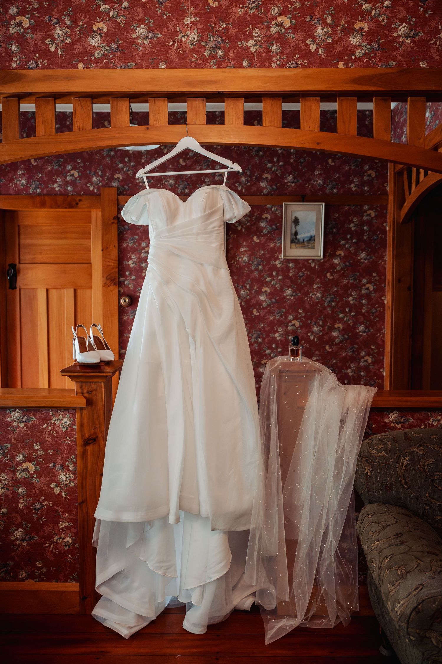 White off-shoulder wedding dress hanging in a vintage room with floral wallpaper, bridal heels and veil nearby.