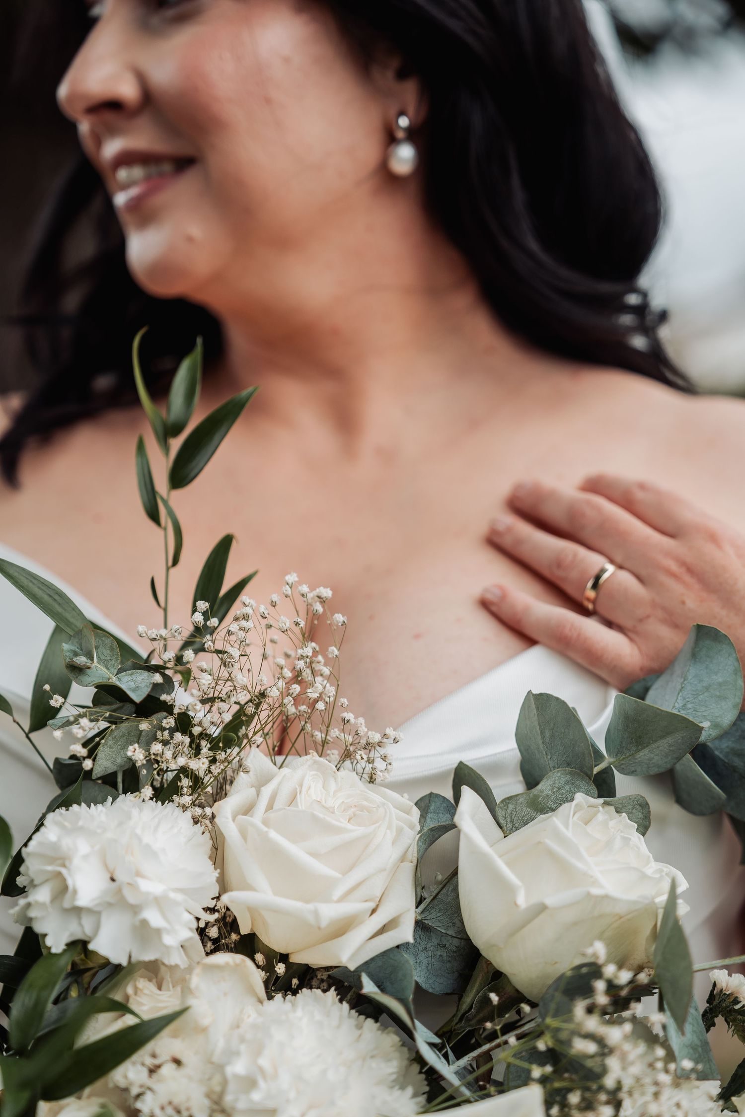 Close-up of a bride holding white roses and eucalyptus bouquet with a gold wedding ring on her finger.