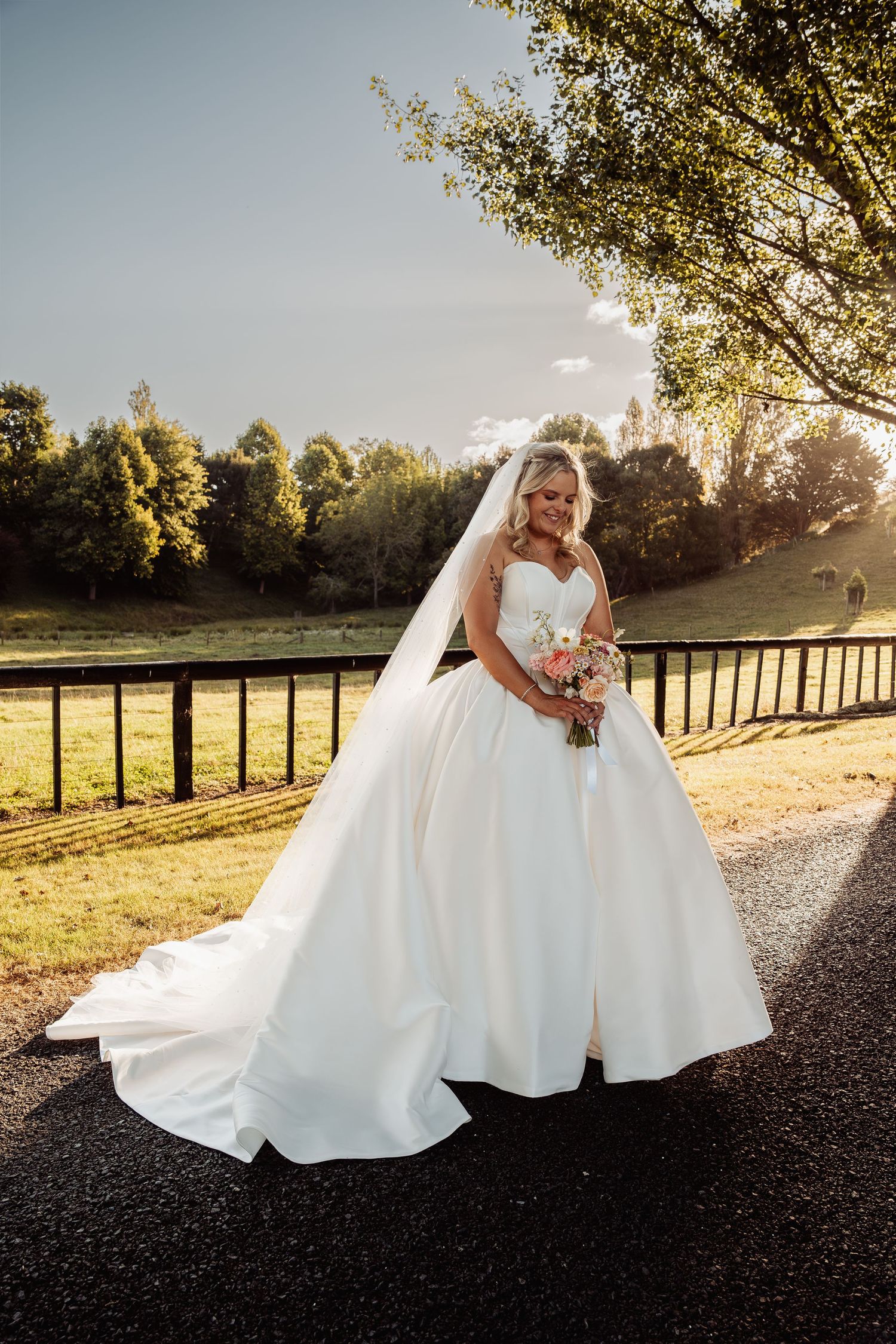 Bride in white ballgown with veil poses on path in golden sunset light.