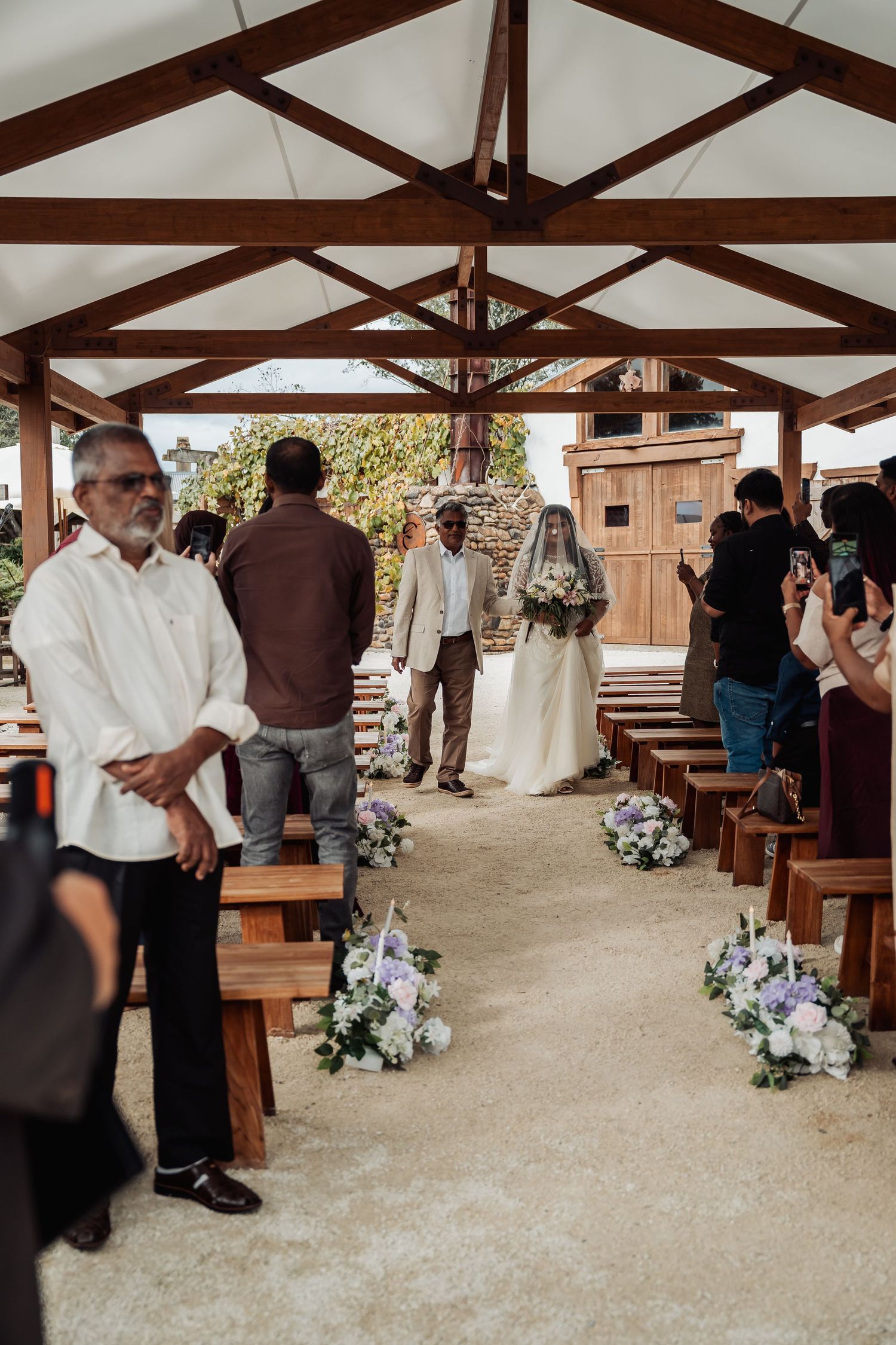 Bride walking down outdoor aisle with guests seated on wooden pews at wedding.