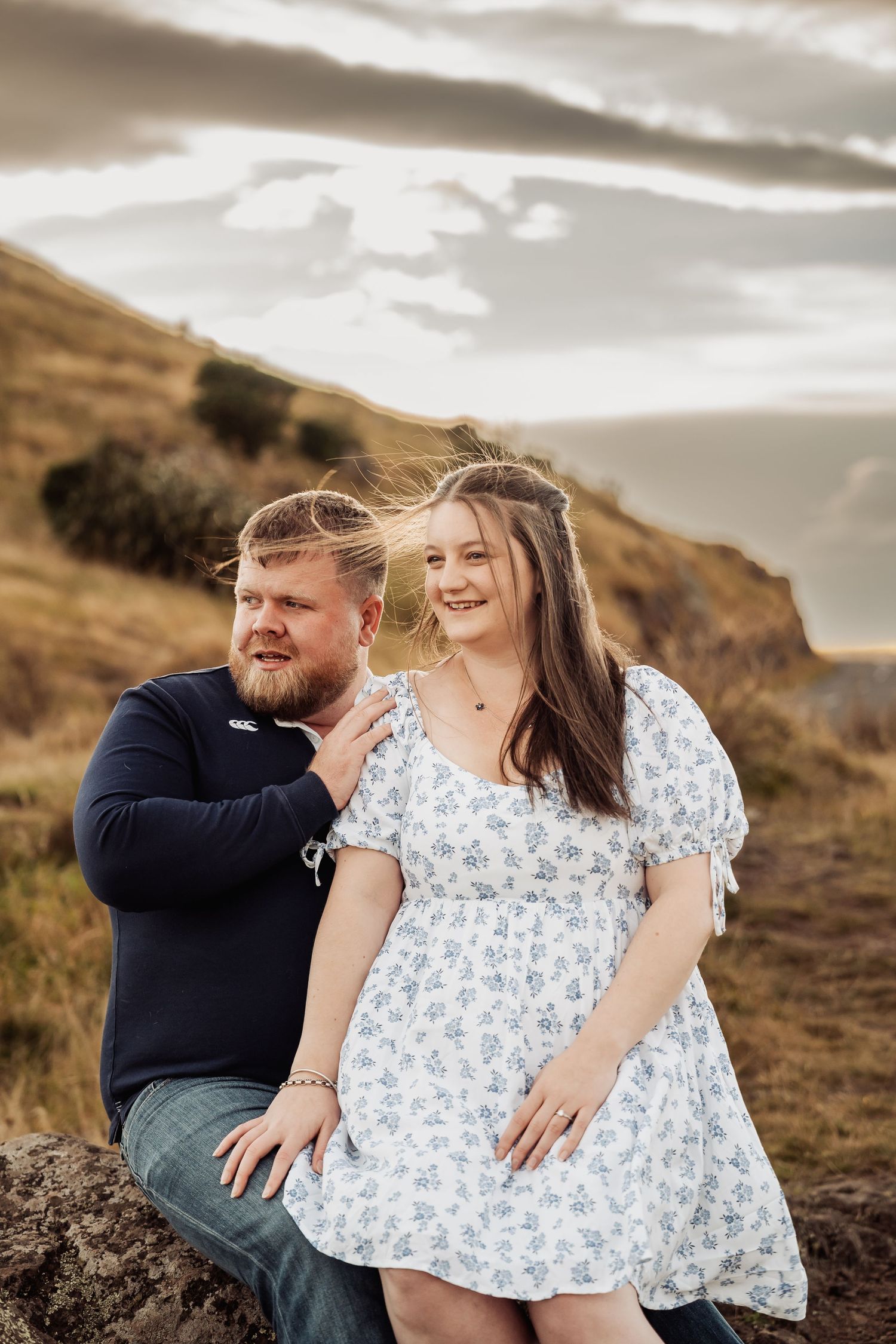 Couple sitting together on rocks with golden hills and dramatic cloudy sky behind them.