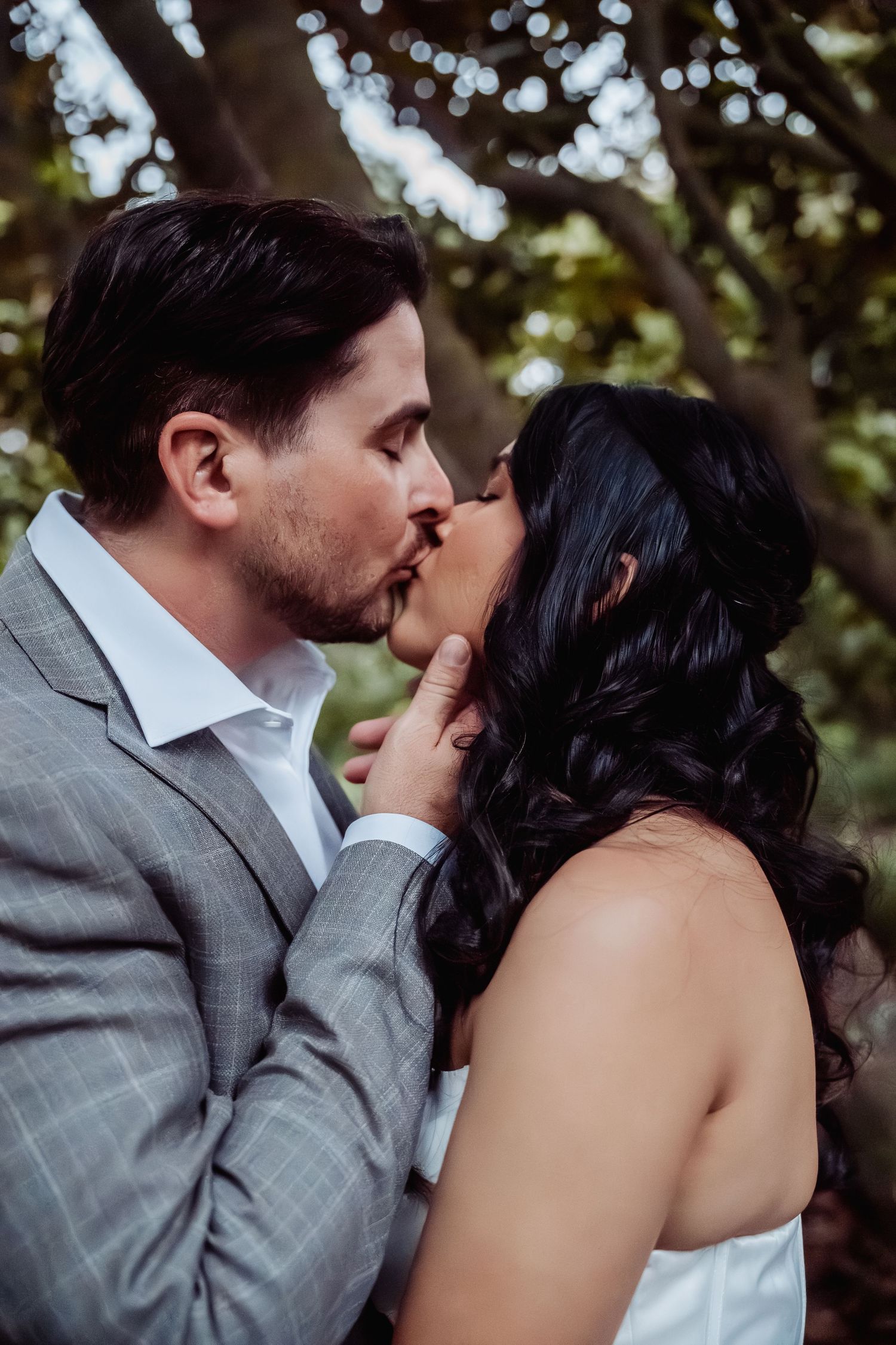 Groom tenderly cups bride's face as they share a romantic kiss beneath trees at their wedding.