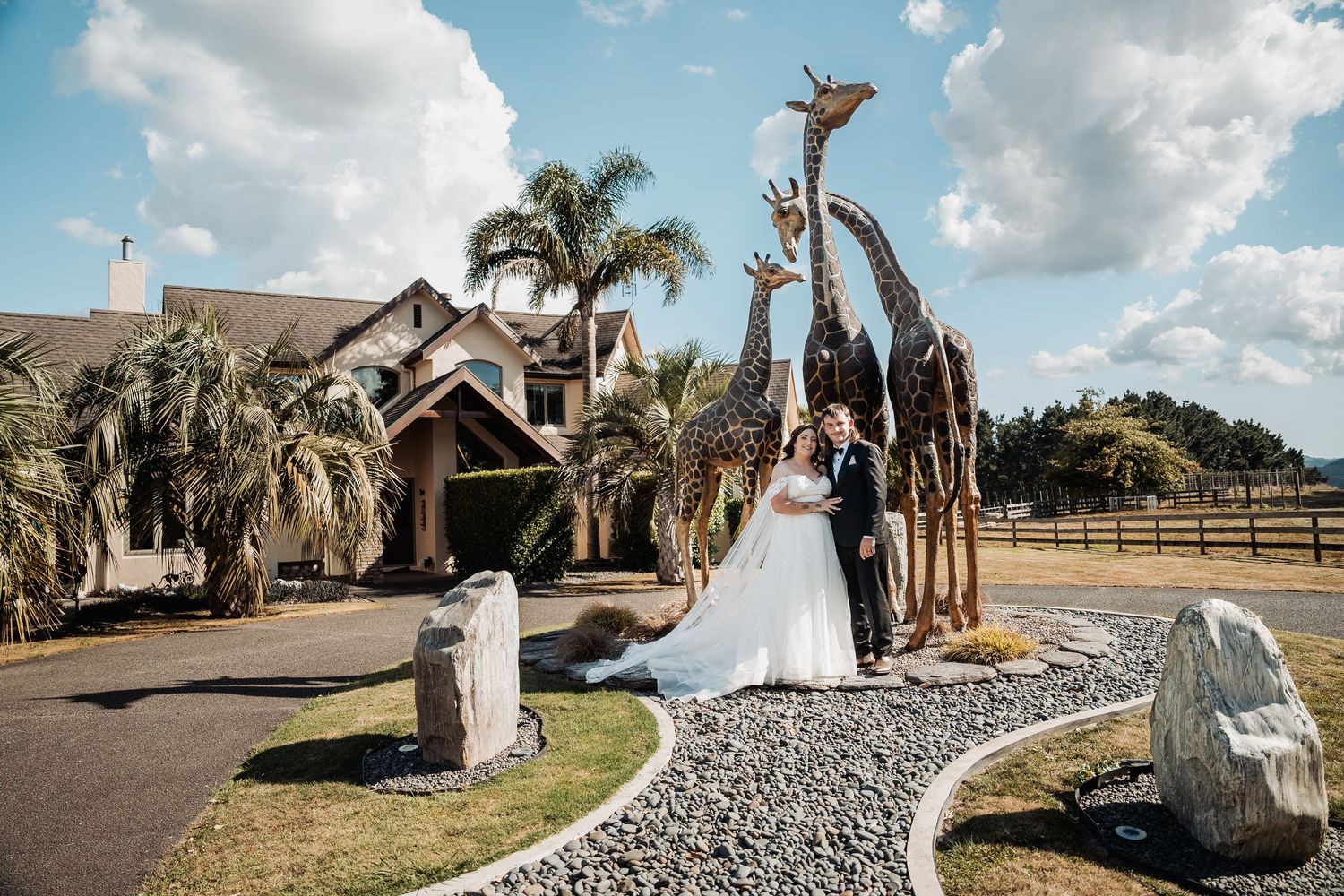 Bride and groom pose beside tall giraffe sculptures outside elegant venue with palm trees.