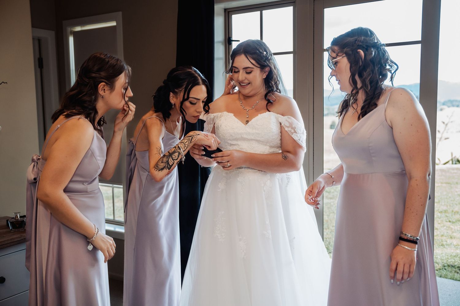 Bride in off-shoulder white gown sharing a joyful moment with three bridesmaids in lavender dresses indoors.