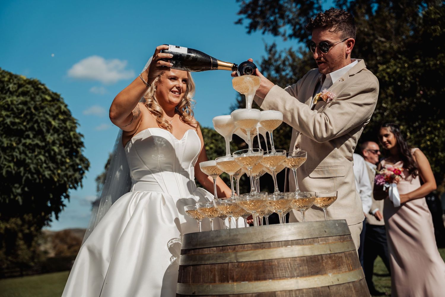 Bride and groom pouring champagne tower on a barrel at outdoor wedding celebration under blue sky.