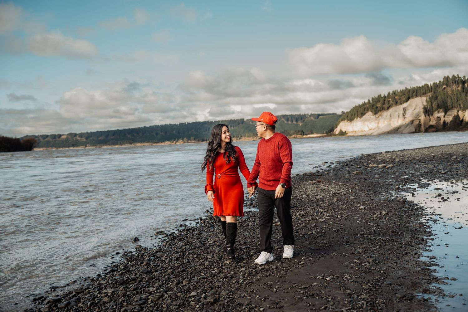 Happy couple in matching red outfits hold hands while walking along a rocky beach shoreline.