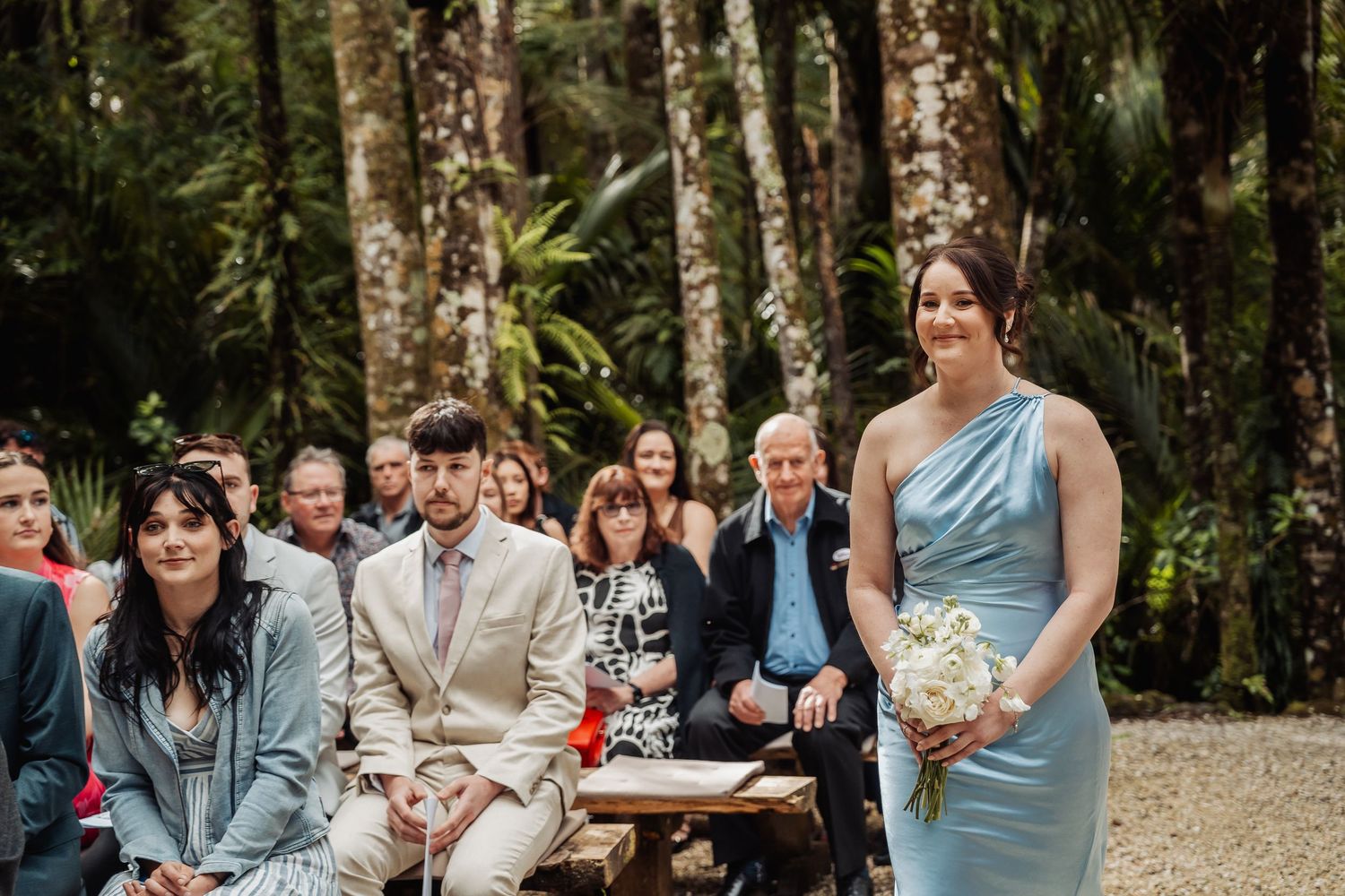 Outdoor wedding ceremony with guests seated in a wooded setting surrounded by tall trees.