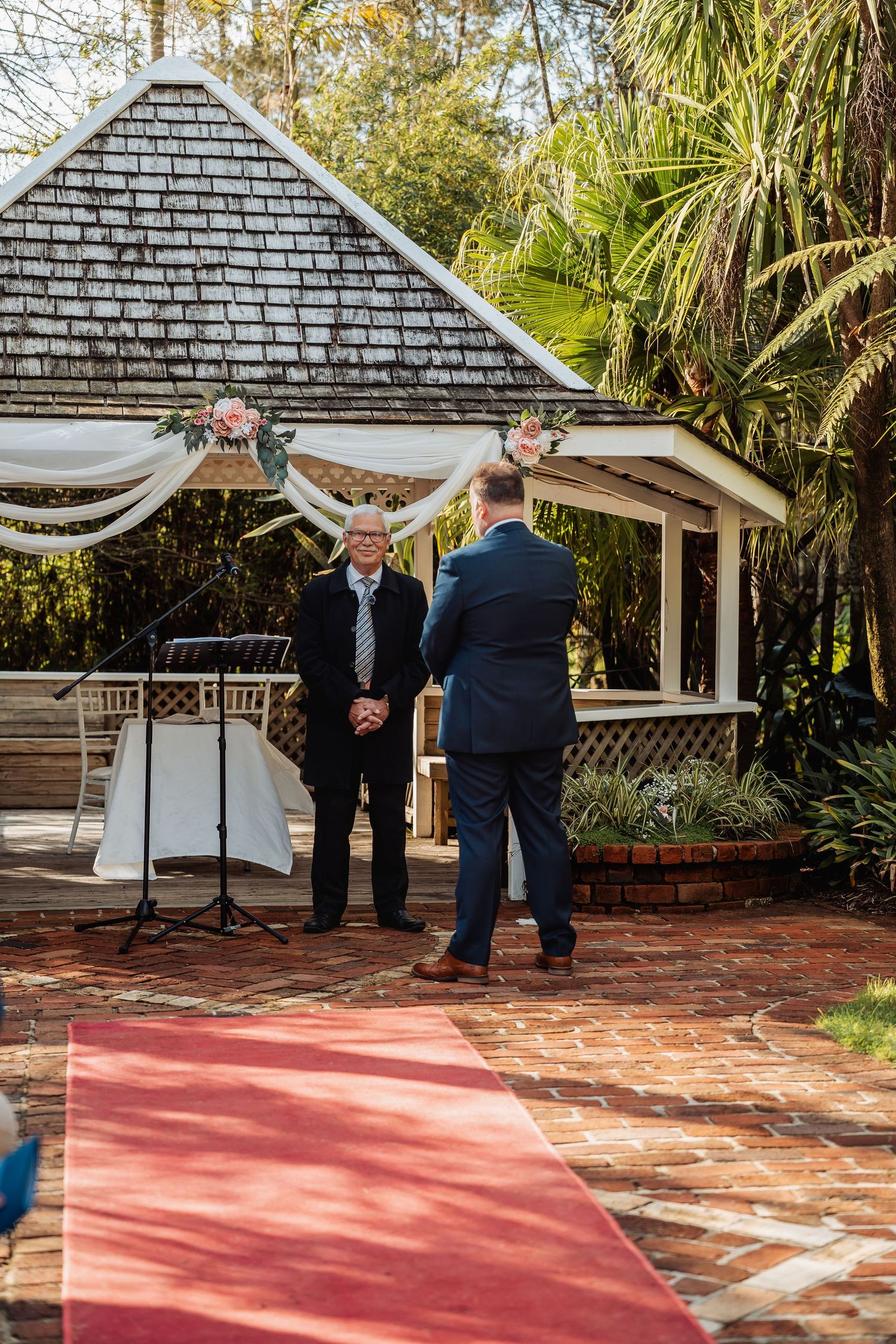 Outdoor wedding gazebo with white draping and brick walkway surrounded by lush tropical landscaping.