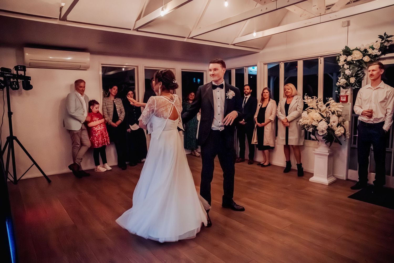 Wedding guests watch as a couple shares their first dance on a wooden dance floor in an elegantly lit reception venue.