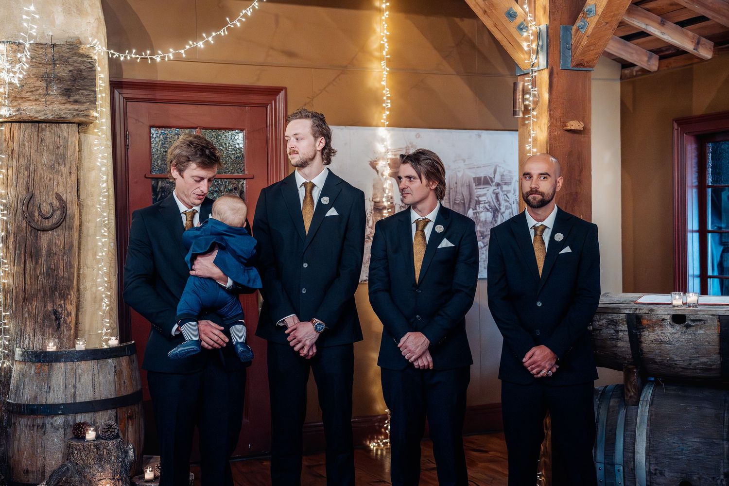 Four people in formal attire stand in a rustic venue with wooden beams and string lights at a wedding ceremony.