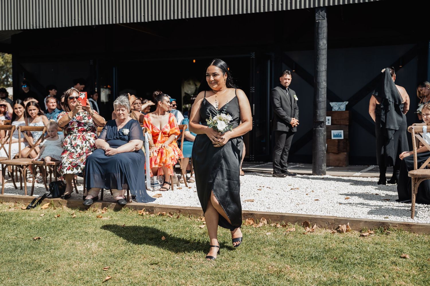 A procession walks along a garden path while guests sit on white chairs during an outdoor ceremony.