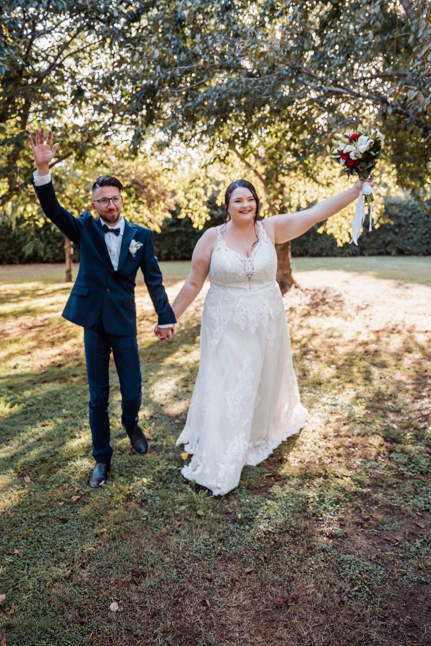 Happy couple raises arms in celebration on their wedding day in sunlit garden.
