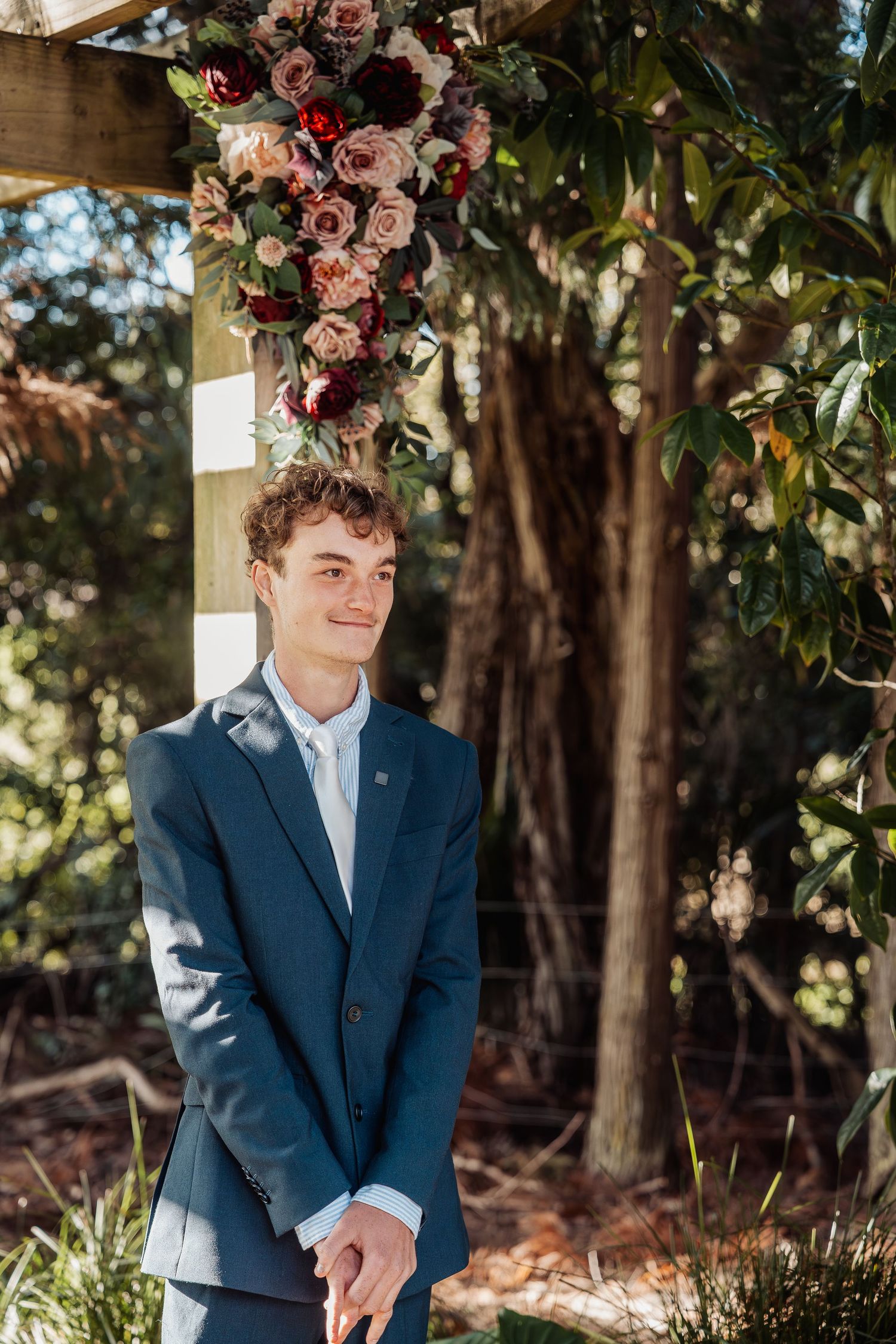 Groom in navy suit standing at floral wedding arch outdoors.