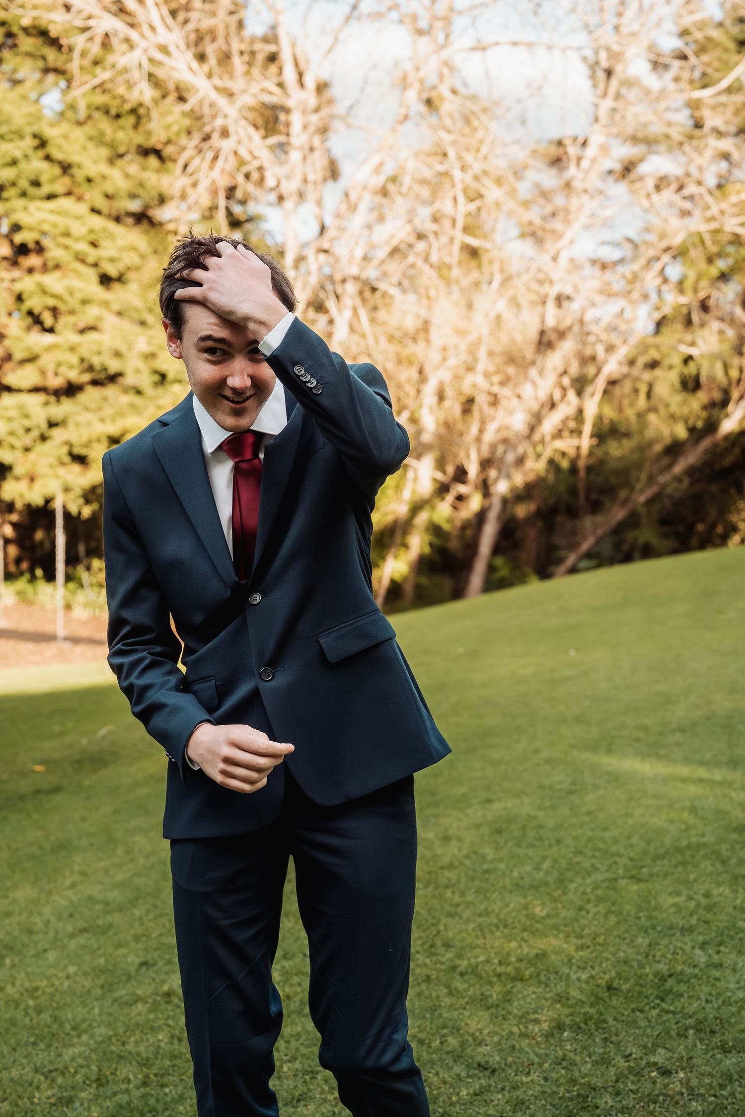 Groom in navy suit and red tie laughing while running hand through hair on green lawn.