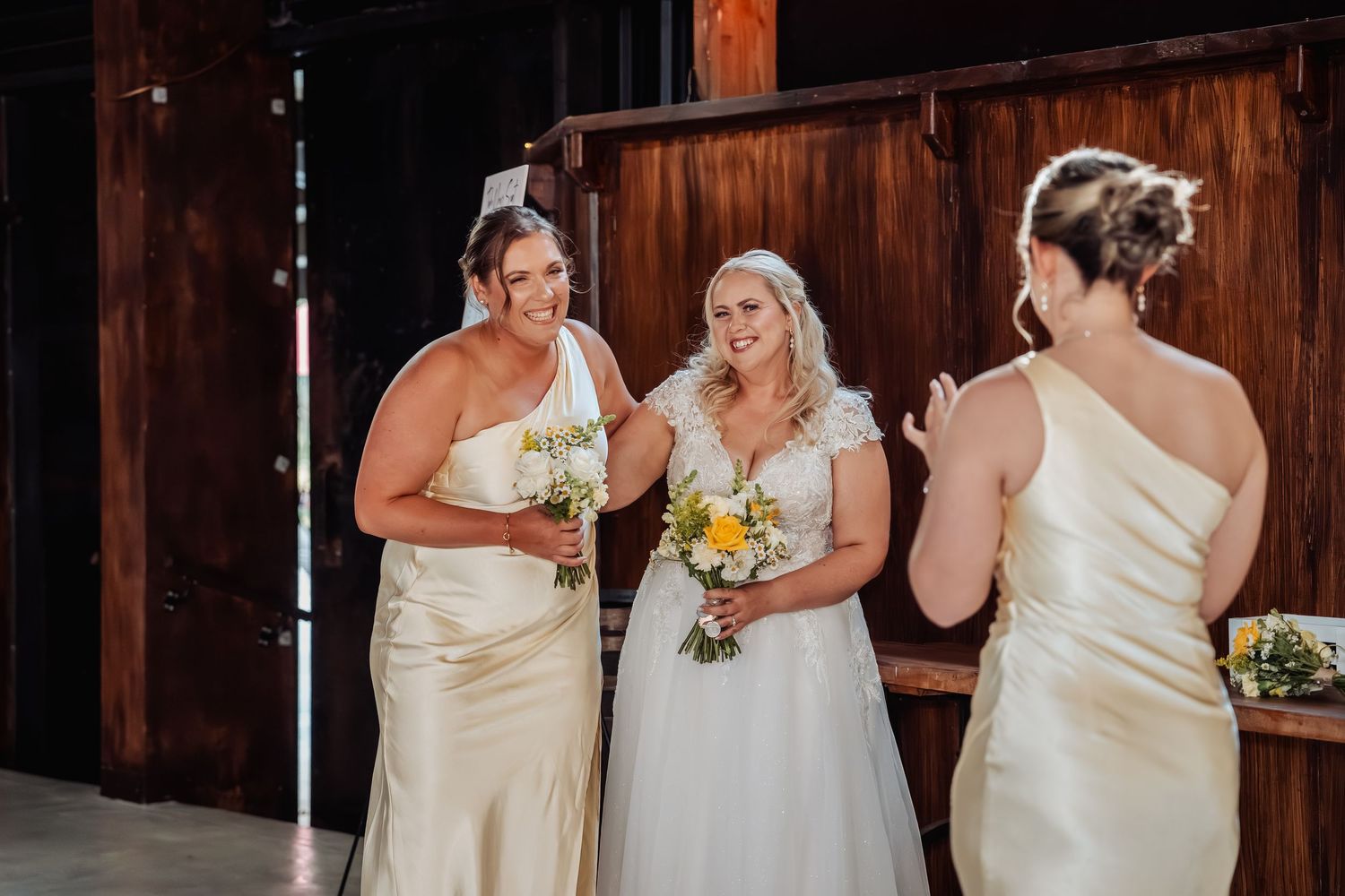 Bride in white gown smiles with two bridesmaids in ivory dresses holding yellow bouquets in rustic barn venue.