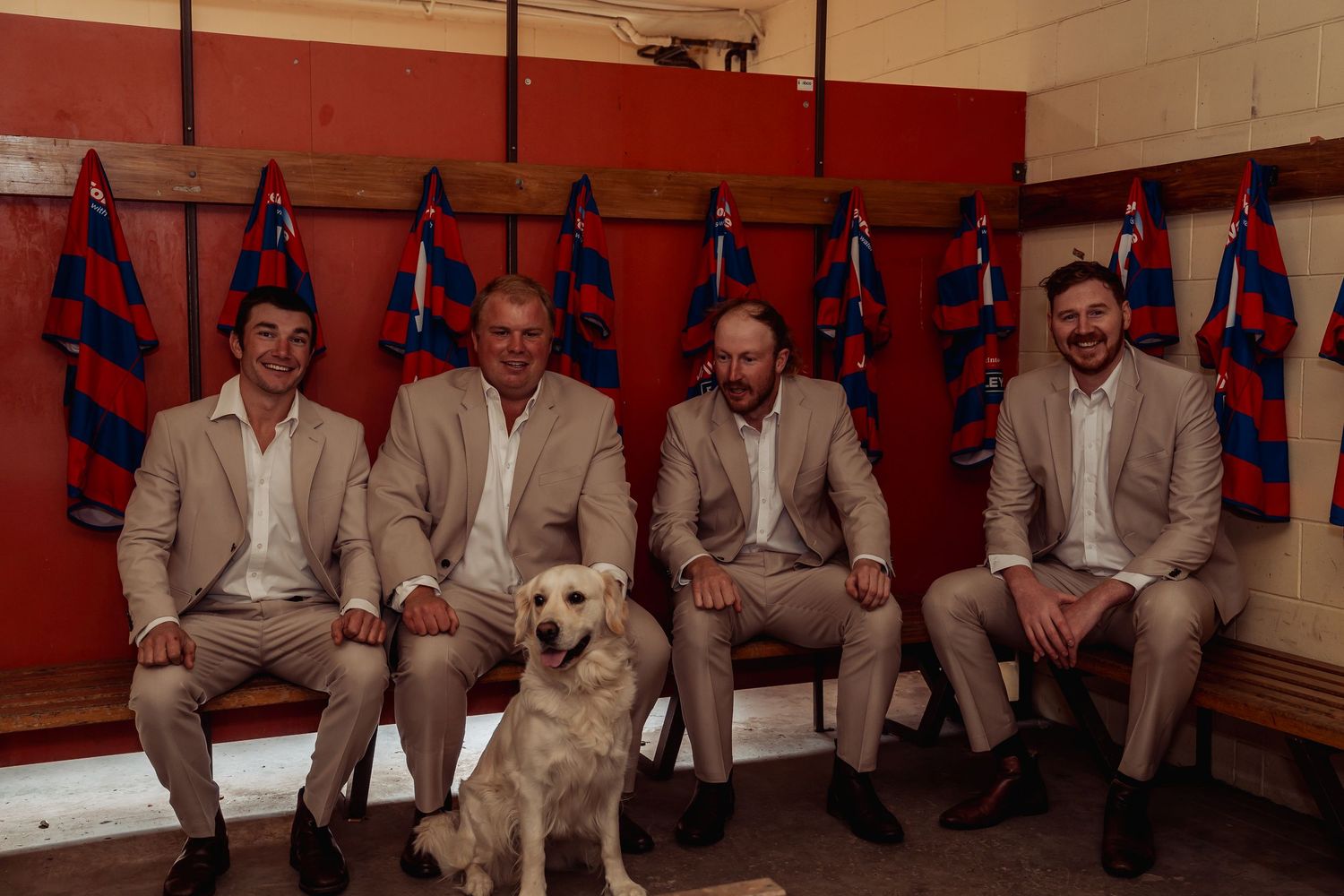Four men in matching tan suits sitting on a bench in a sports locker room with a white Labrador dog.