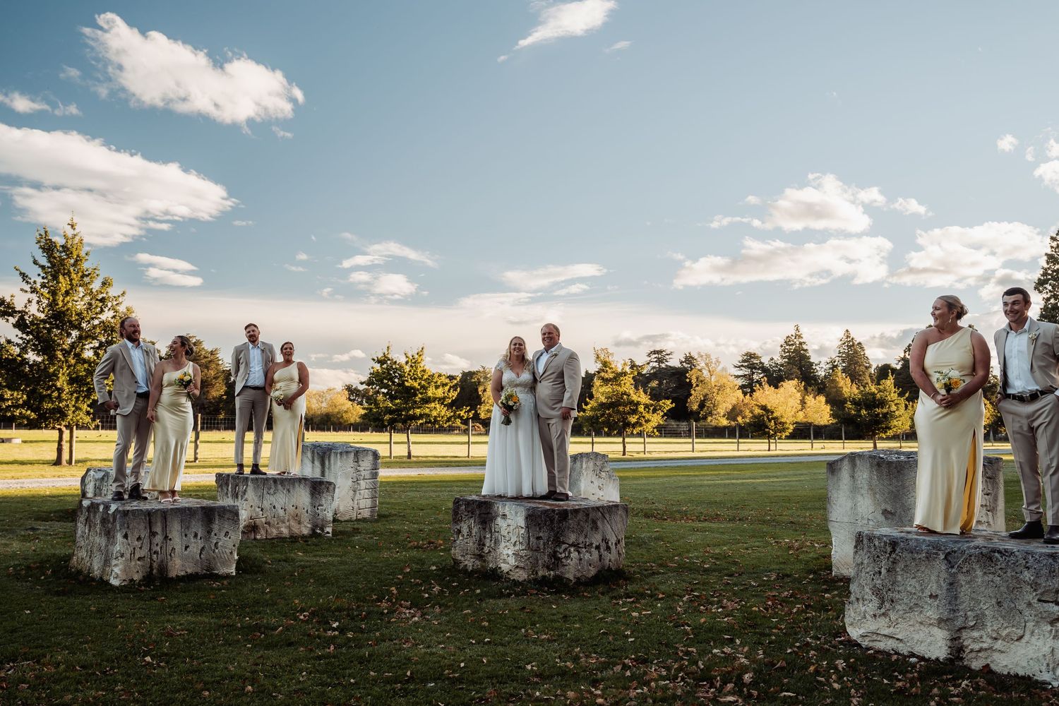 Wedding party stands on stone pedestals in golden hour sunlight at scenic outdoor venue.