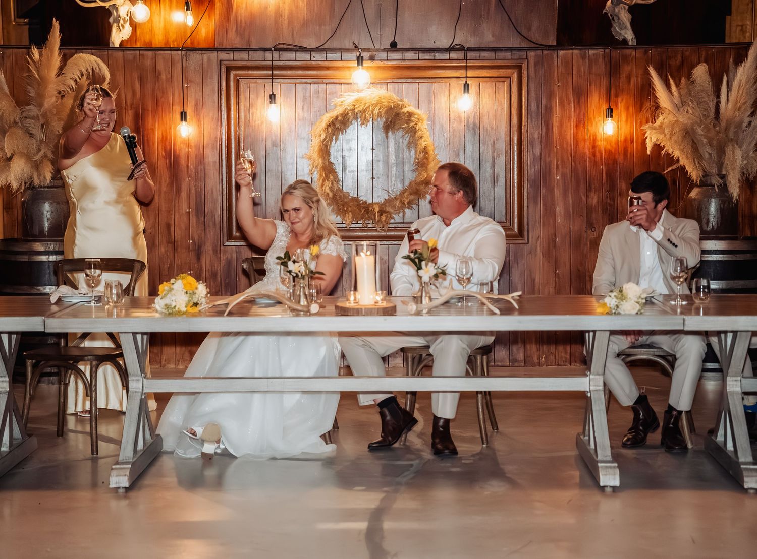 Bride and groom sit at sweetheart table in rustic barn venue as a guest raises a toast during reception.